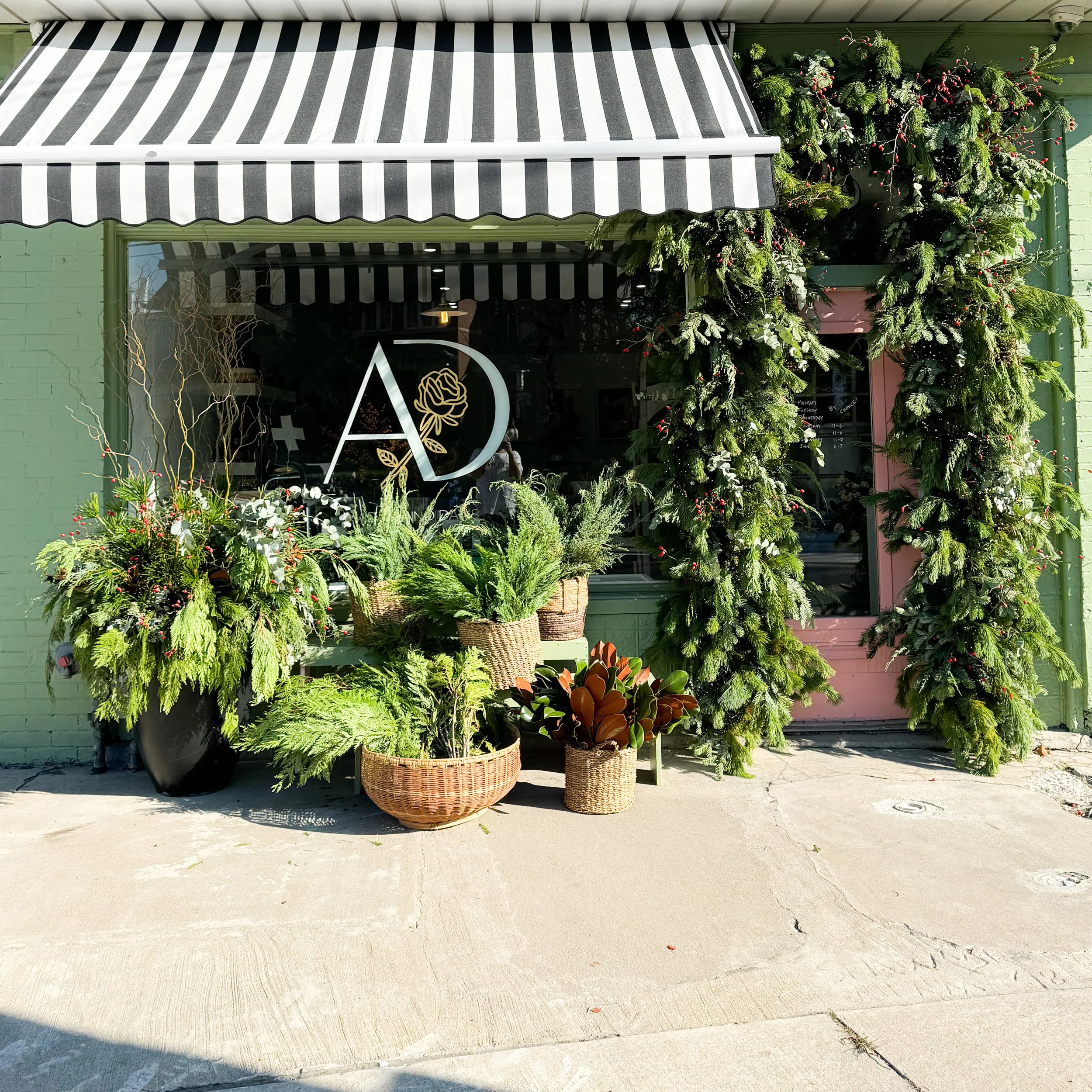 Exterior shop window decorated with lush green plants and wreaths, with a striped awning above and a pink door to the side.