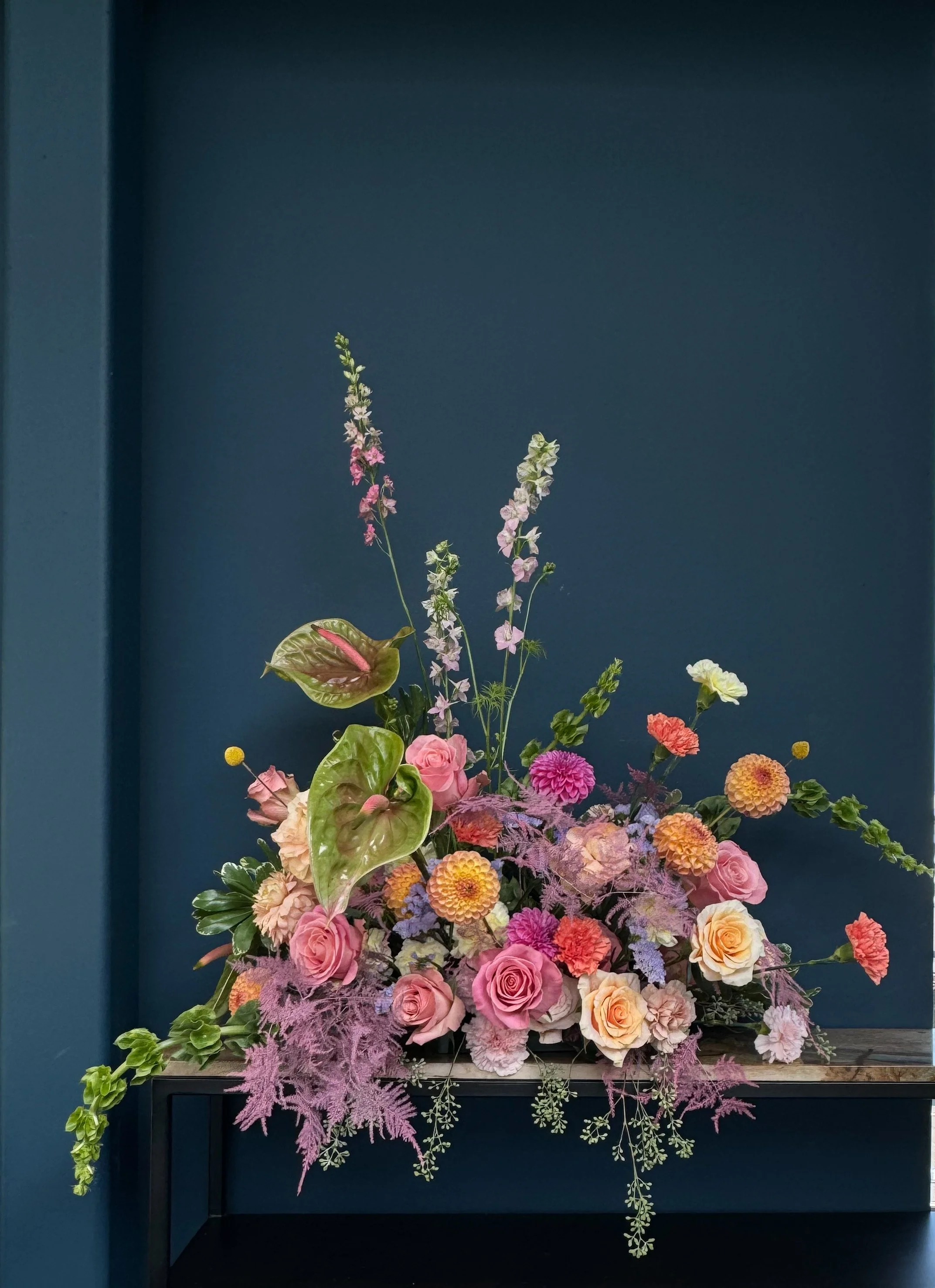 A vibrant arrangement of pink and peach roses, purple dahlias, and assorted greenery and filler flowers displayed on a black table against a dark blue wall.