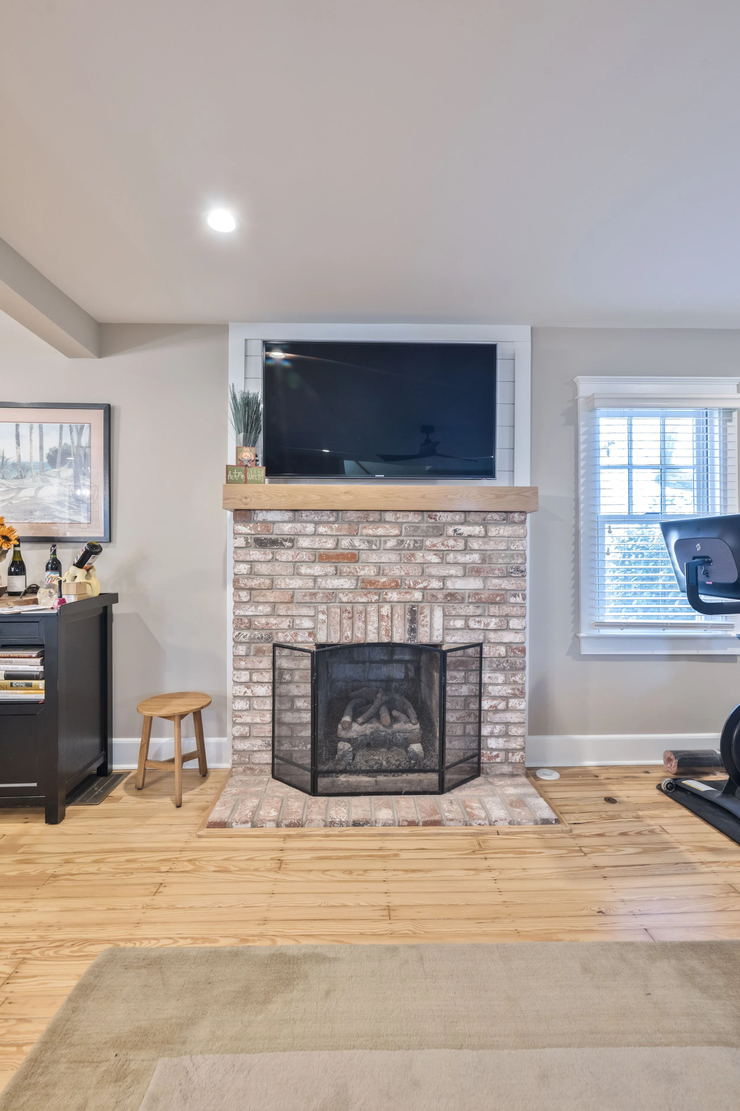 Living room with brick fireplace and mounted flat-screen TV above, window with white blinds on right, and exercise equipment on the right side.