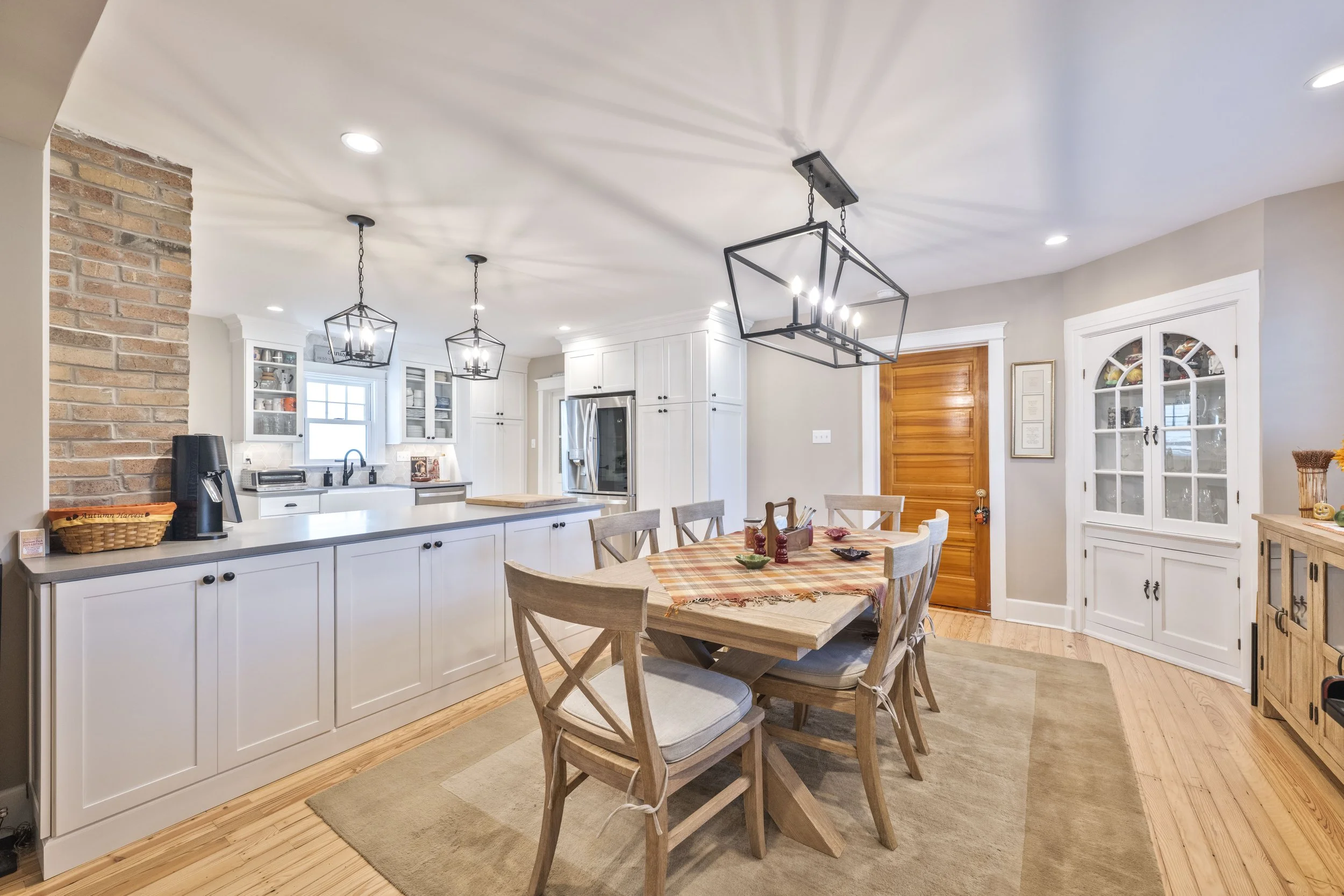 Kitchen and dining area with wooden flooring, white cabinets, a brick wall, and a dining table with six chairs, covered with a plaid tablecloth. Lighting fixtures hang from the ceiling.