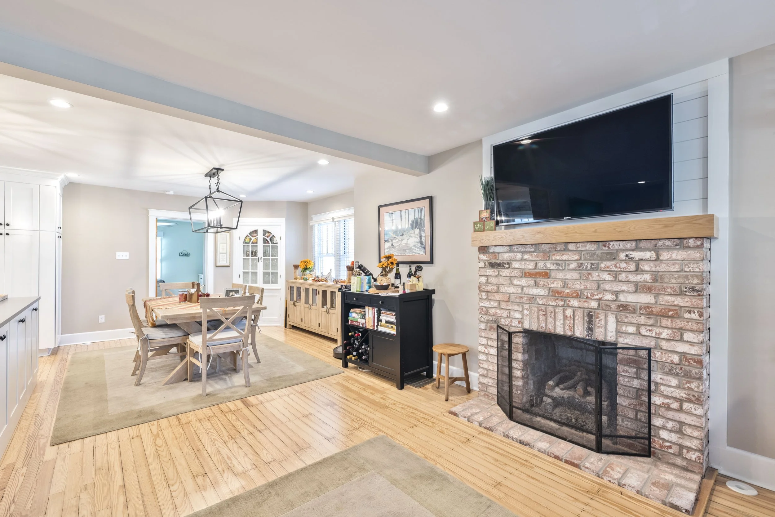 Living room and dining area with hardwood floors, brick fireplace with a mounted TV, dining table with six chairs, sideboards, and windows with blinds.