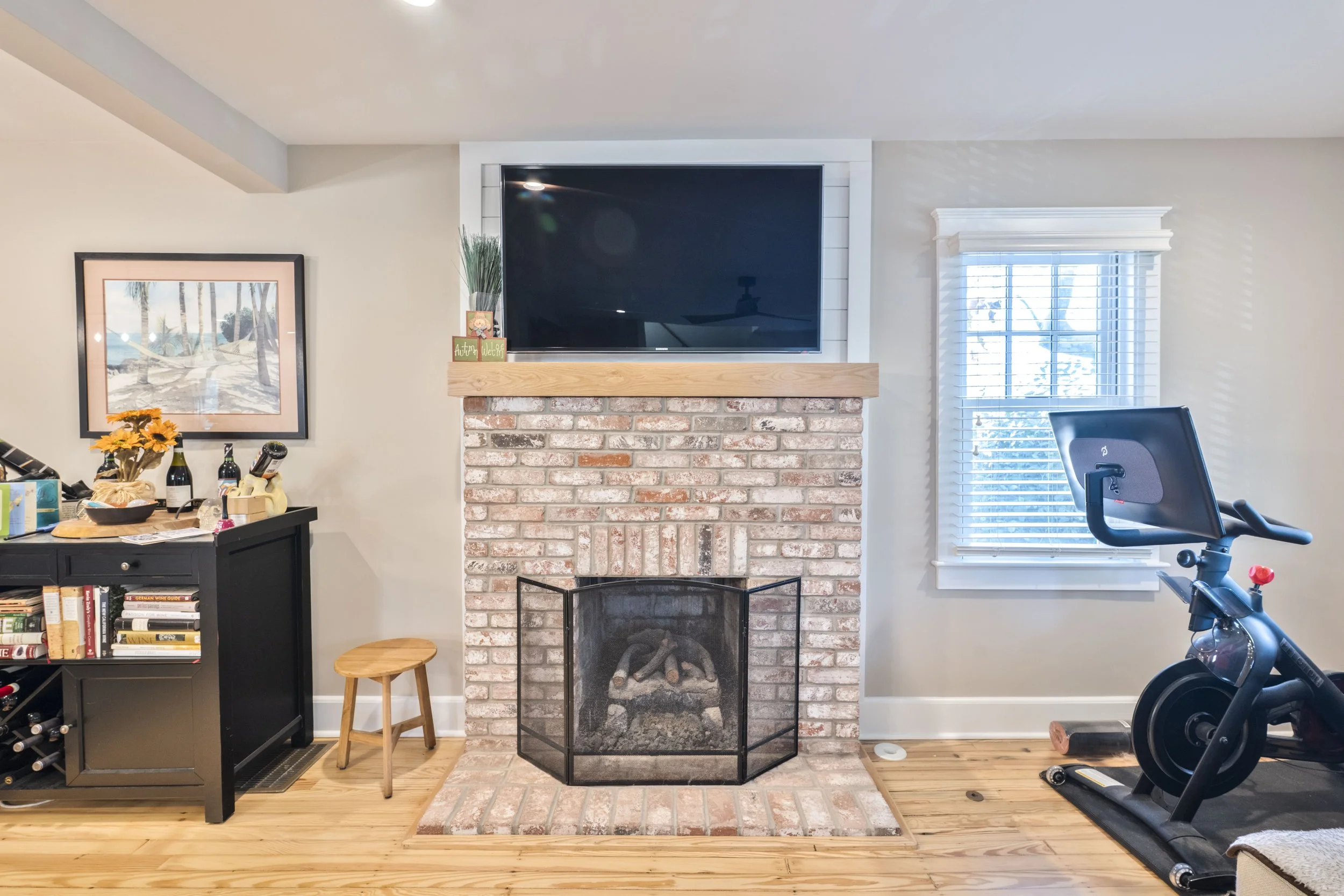 A living room with a brick fireplace topped by a flat-screen TV, a black bookshelf with bottles and books, a small wooden stool, a window with white blinds, and a stationary exercise bike nearby.