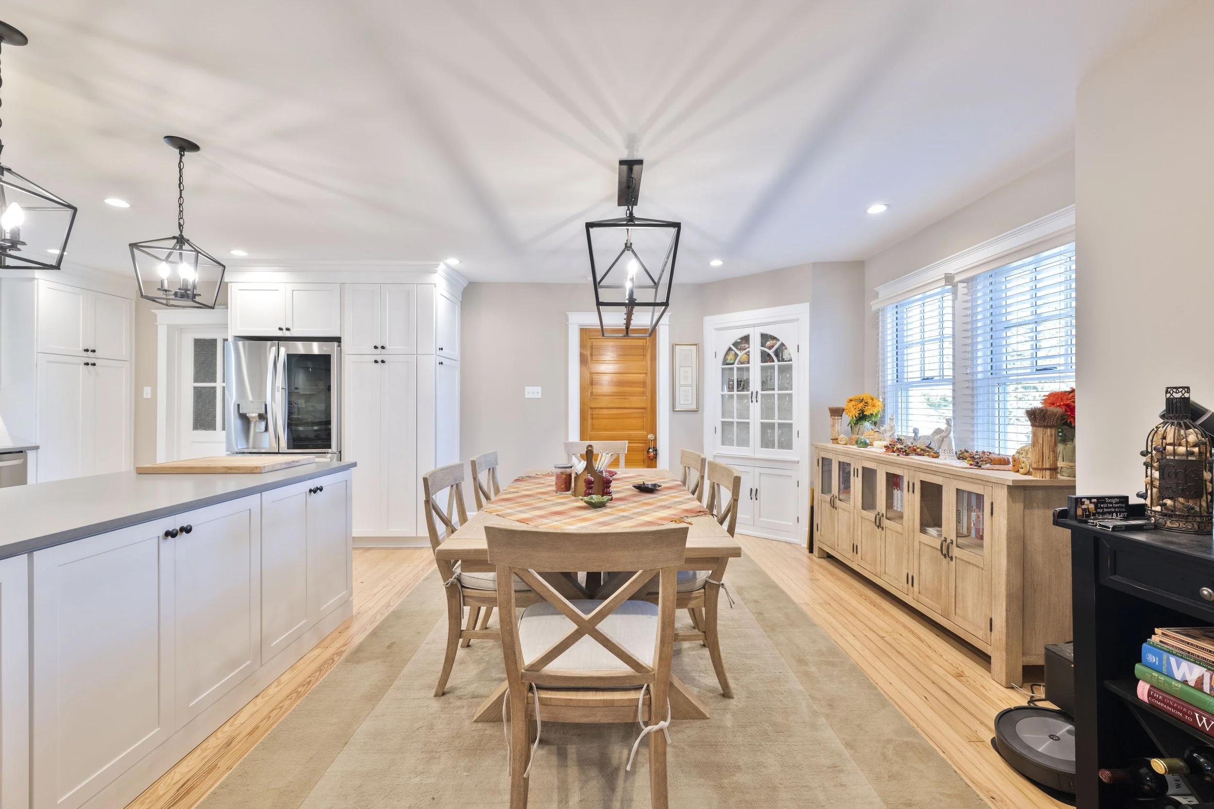 A spacious kitchen and dining area with white cabinets, a wooden dining table with chairs, and large windows with white blinds, decorated with flowers and fall-themed ornaments.