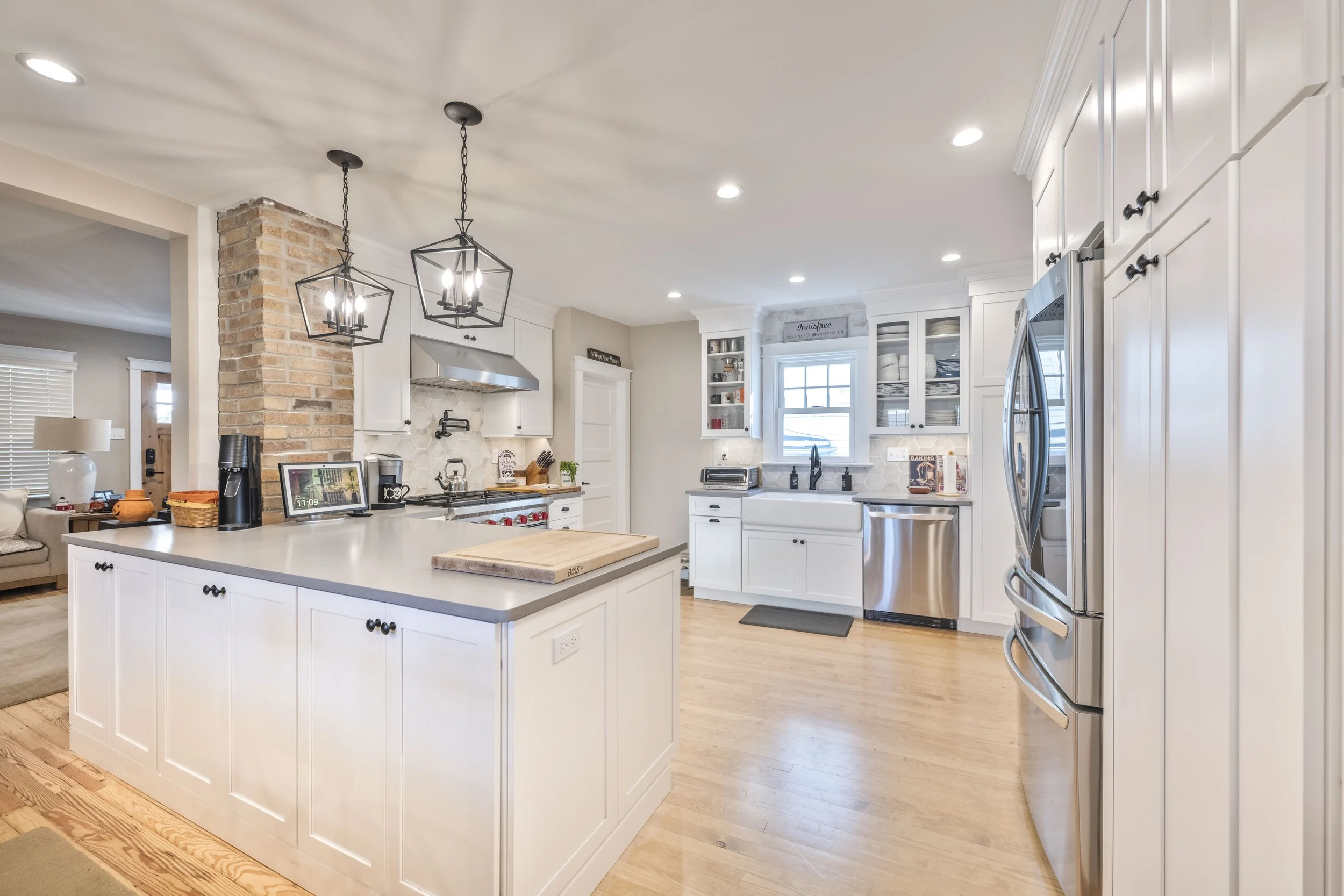 Bright kitchen with white cabinets, stainless steel appliances, a central island with a cutting board, open shelving, and a brick accent wall.