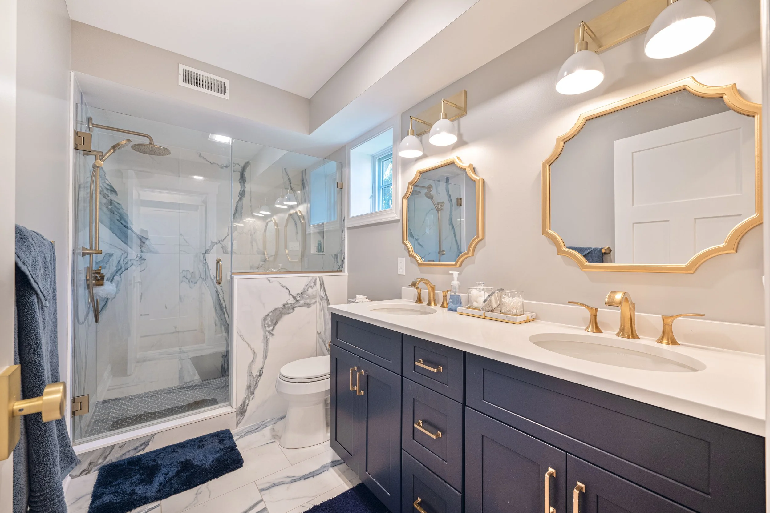 Modern bathroom with double vanity with navy blue cabinets, white countertop, and gold fixtures. Two ornate gold-framed mirrors above each sink, three wall-mounted lights, a glass-enclosed shower with marble tiles, and a small window with natural light.