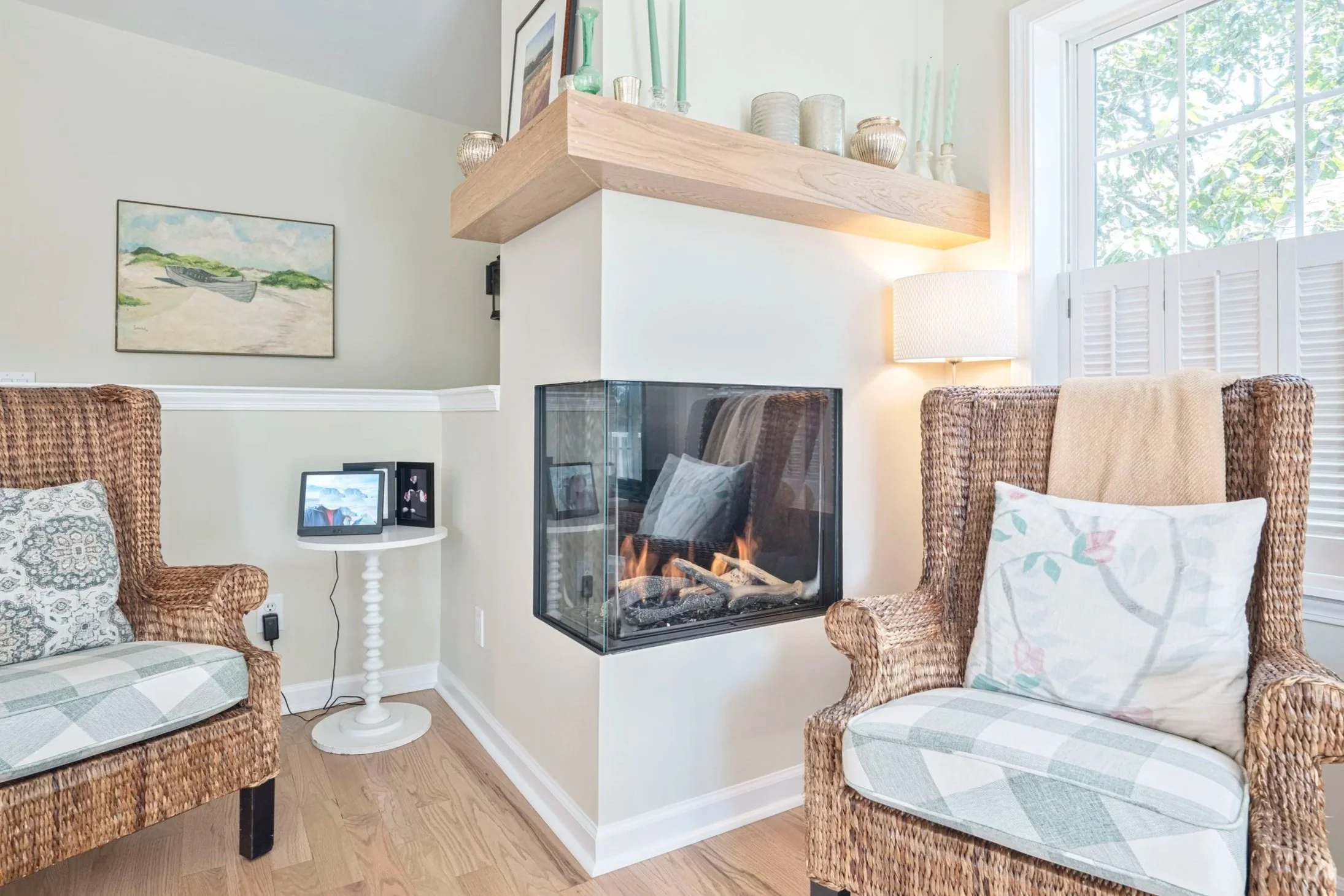 Living room with wicker armchairs, a modern fireplace, a window with white shutters, a side table with a digital photo frame, and decorative items on a wooden shelf