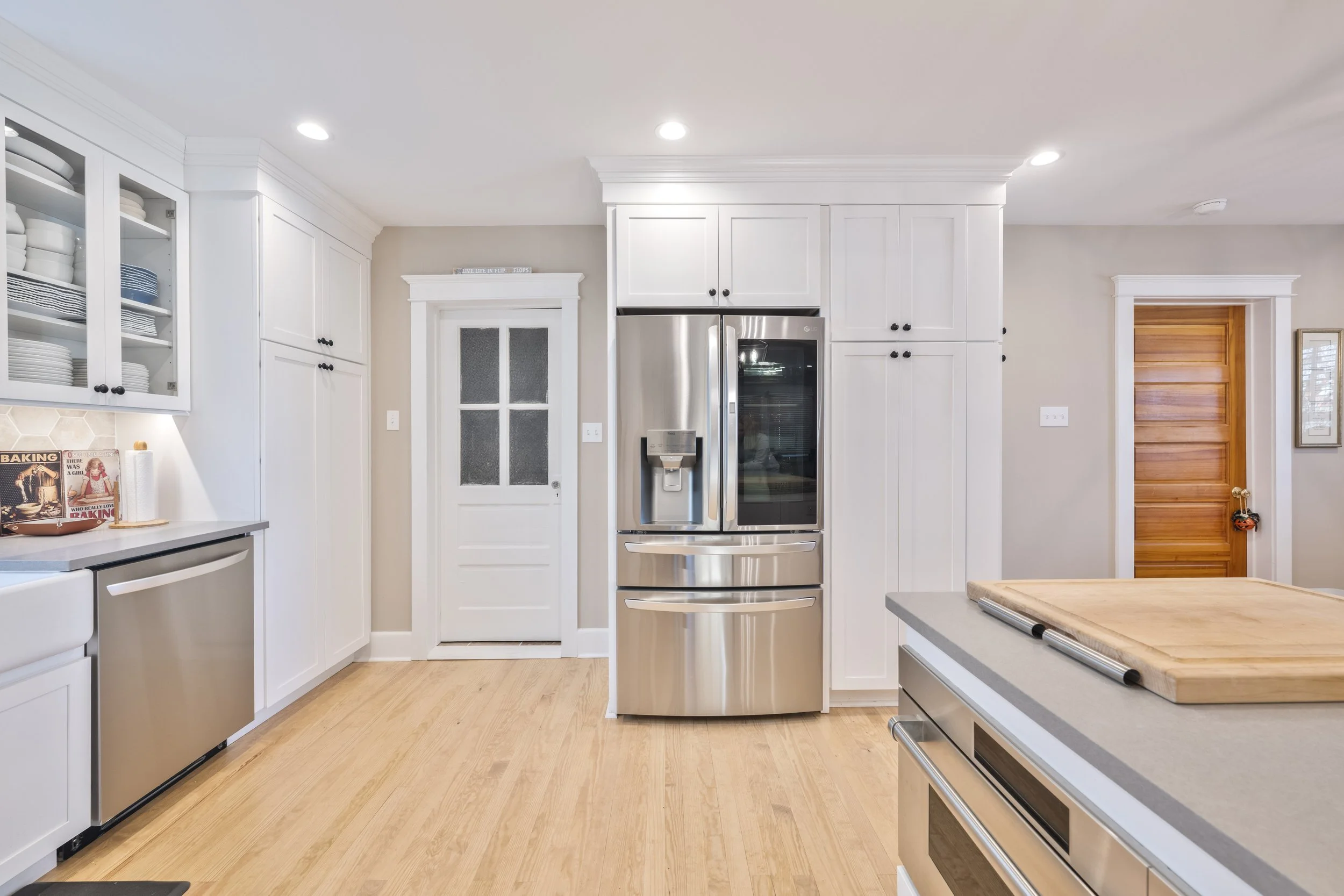 Modern kitchen with stainless steel refrigerator, white cabinets, wooden floor, and a wooden door.