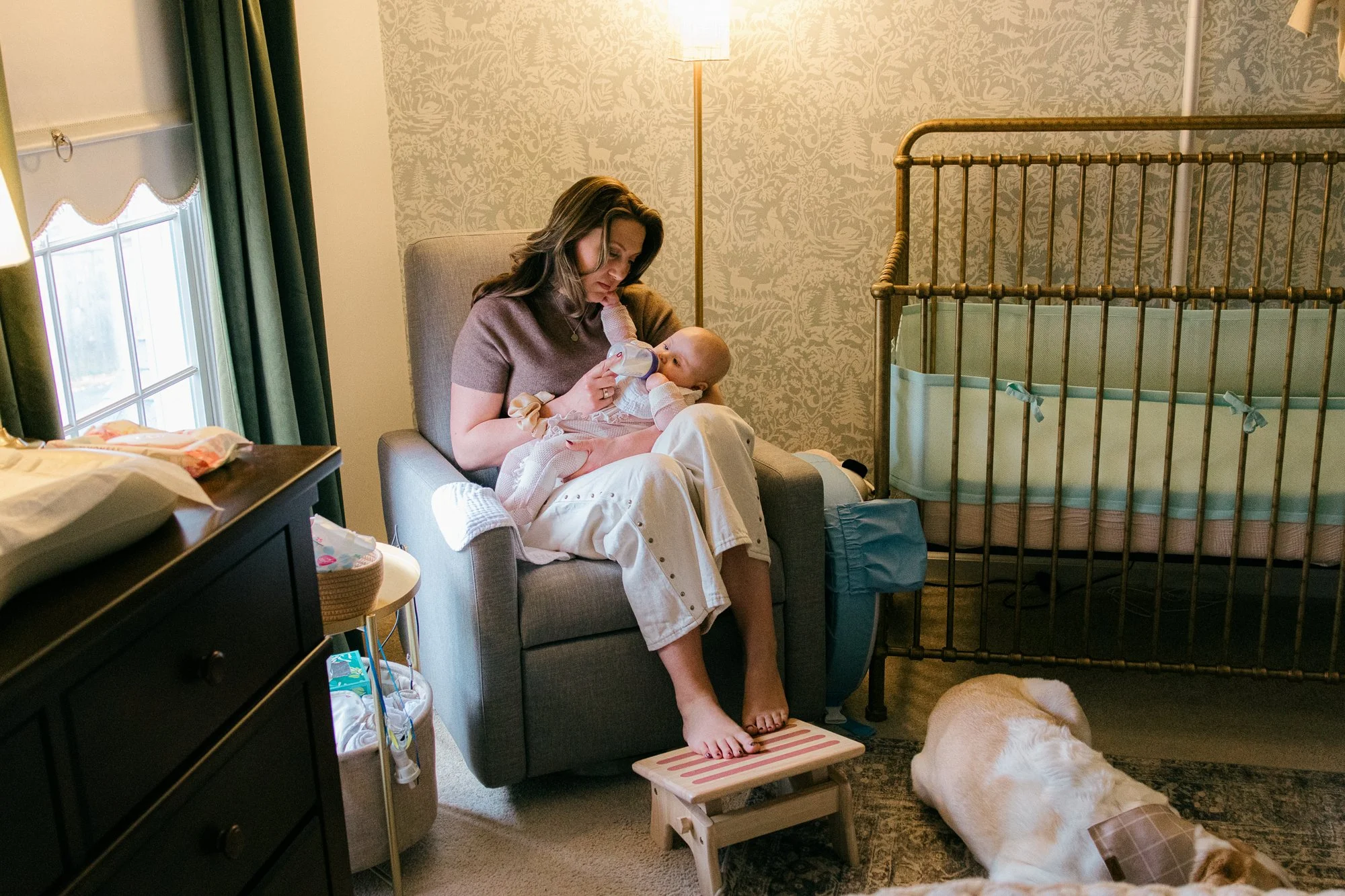  Documentary family photo of a mother bottle-feeding her baby in the nursery while the family dog rests nearby in their Sterling, Virginia home. 