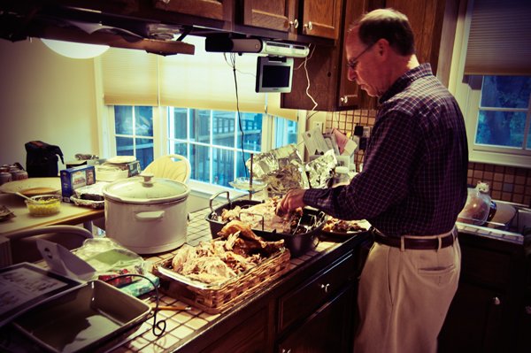 A man in a plaid shirt carves meat in a home kitchen during Passover meal preparation, captured candidly behind the scenes by Mel Usry Photography Northern Virginia.