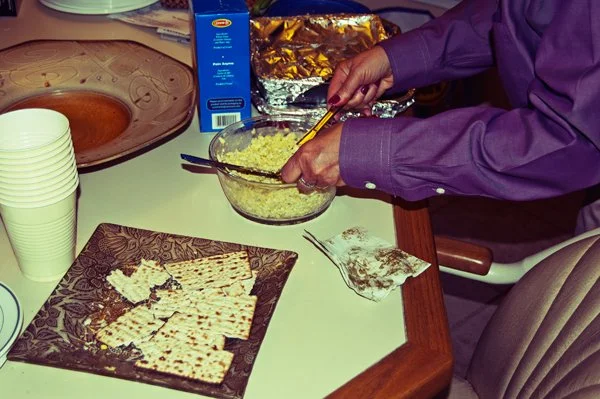 Hands serving egg salad from a bowl beside a plate of broken matza during a Passover seder gathering, documented candidly by Mel Usry Photography Northern Virginia.