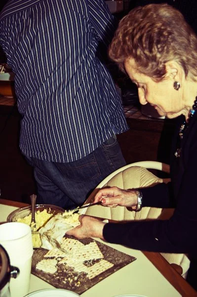 An older woman leans forward to serve herself from a plate of matza and egg salad at a Passover gathering, captured candidly by documentary family photographer Mel Usry Photography.