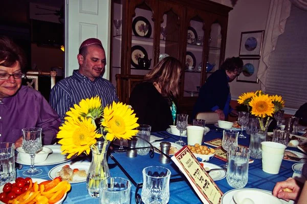 Family members seated candidly around a Passover seder table with sunflowers, crystal glasses, and haggadahs, documented by documentary family photographer Mel Usry Photography in Northern Virginia.