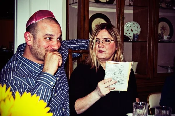 A man wearing a kippah laughs with a woman holding a haggadah at the Passover seder table, an unposed candid moment captured by documentary family photographer Mel Usry Photography.