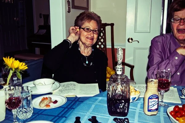An older woman smiles at the Passover seder table surrounded by crystal glasses and traditional Passover foods, captured candidly during a multi-generational family gathering by Mel Usry Photography.