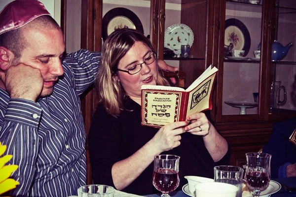 A woman reads aloud from a Passover haggadah while a man wearing a kippah leans in beside her, captured candidly by documentary family photographer Mel Usry Photography in Northern Virginia.