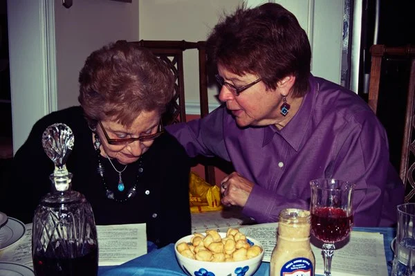 Two women lean together looking at a haggadah at the Passover seder table, a candid multi-generational family moment documented by Mel Usry Photography Northern Virginia.