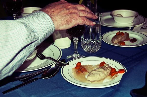 An older man's hand rests near a plate of gefilte fish and crystal glassware at the Passover seder table, captured candidly by documentary family photographer Mel Usry Photography.