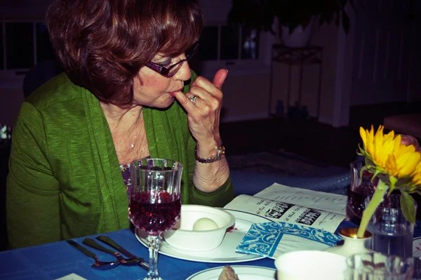 A woman in a green cardigan licks her finger while seated at the Passover seder table with wine and sunflowers nearby, captured candidly by Mel Usry Photography Northern Virginia.