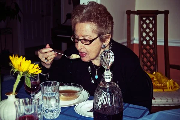 An older woman leans in to eat from a bowl of matzo ball soup at the Passover seder table, captured candidly during a multi-generational family gathering by Mel Usry Photography Northern Virginia.