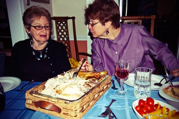 Two women sit together at the Passover seder table with a basket of chicken between them, a candid multi-generational moment documented by Mel Usry Photography Northern Virginia.
