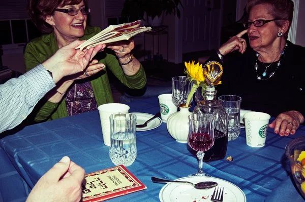 A woman laughs while fanning haggadahs across the Passover seder table as an older woman smiles beside her, a candid joyful moment captured by Mel Usry Photography Northern Virginia.
