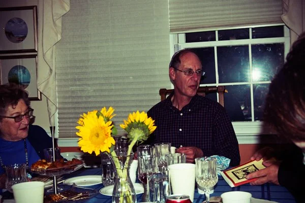 A man wearing a kippah speaks at the Passover seder table beside sunflower centerpieces, captured candidly during a multi-generational family gathering by Mel Usry Photography Northern Virginia.