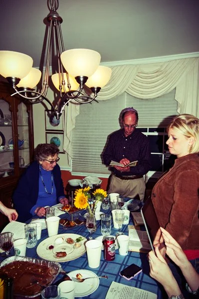 A multi-generational family gathers around the Passover seder table as a man reads from a haggadah while standing, documented candidly by documentary family photographer Mel Usry Photography Virginia.