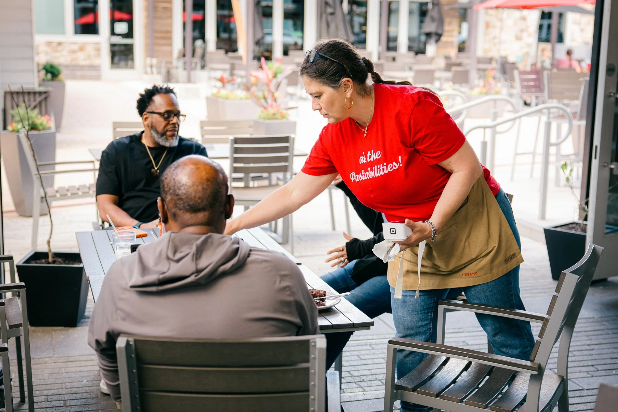 A server in an Urbano red t-shirt and apron serves guests on the outdoor patio, captured candidly during a documentary brand story session by Mel Usry Photography in Sterling, Virginia.