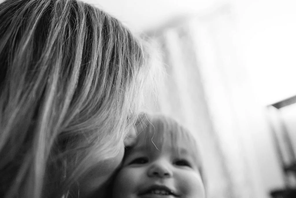 Black and white documentary family photograph of a parent kissing their baby during a quiet, intimate moment at home.
