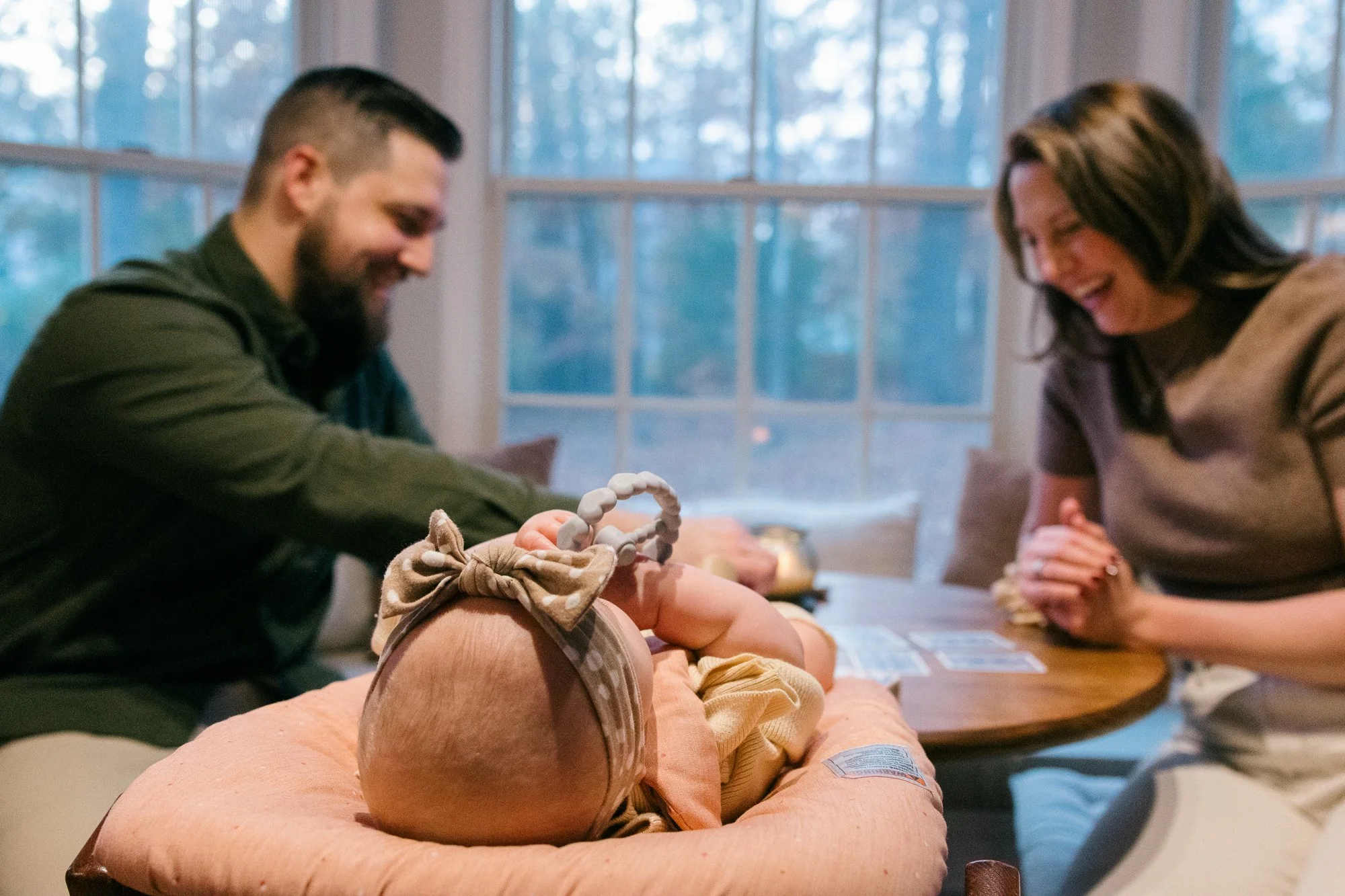 Baby resting in a baby seat beside the table while parents laugh and play a game during an in-home documentary family session in Sterling, Virginia
