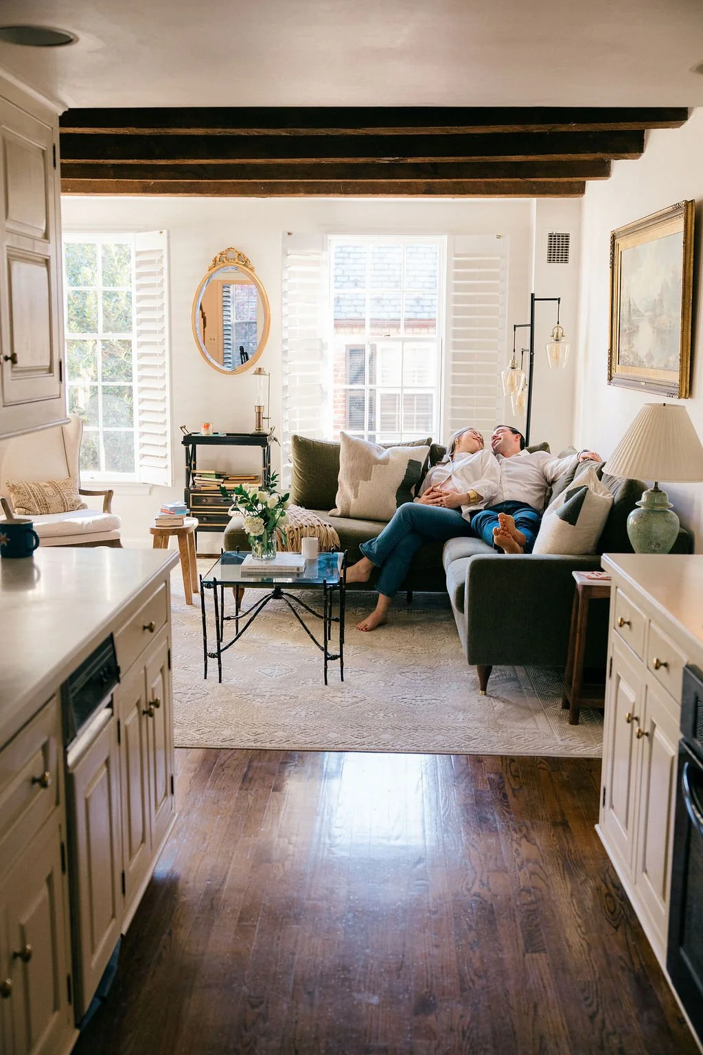 A couple sitting on a gray couch in a bright living room, leaning on each other and laughing. The room has white walls, large windows with shutters, and various decor including a mirror, a framed painting, a table lamp, and a vase with flowers.