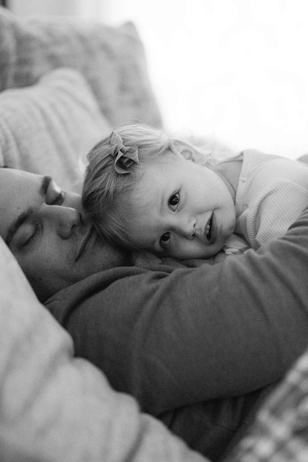 Black and white documentary family photo of a toddler resting their head on a parent during a quiet moment at home.