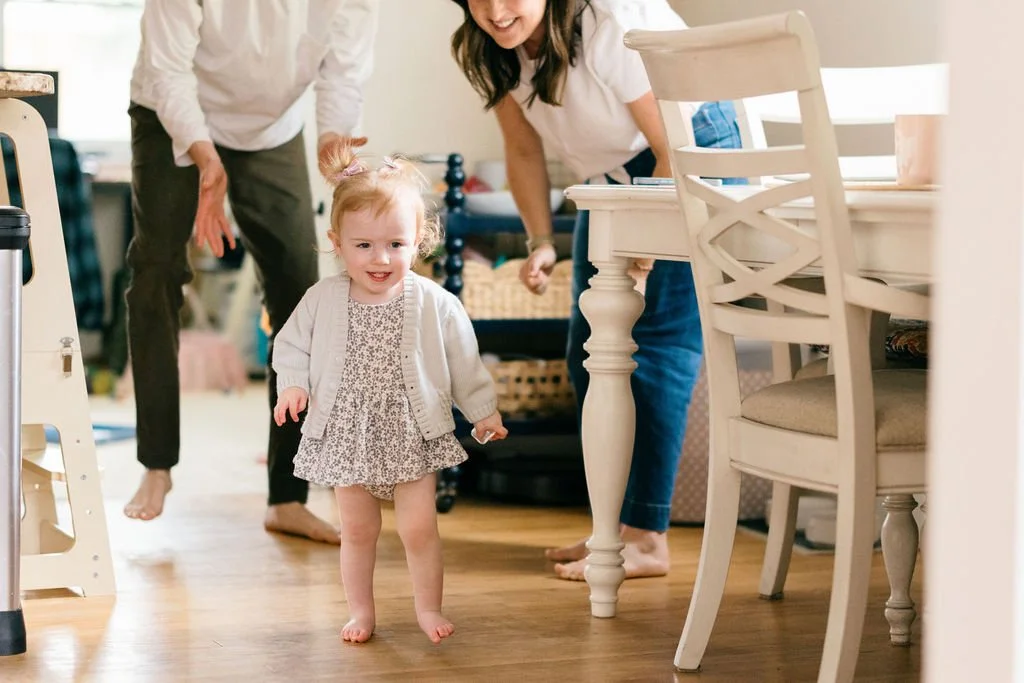 Toddler confidently walking through the living room while parents follow behind during an in-home documentary family session in Alexandria, Virginia.
