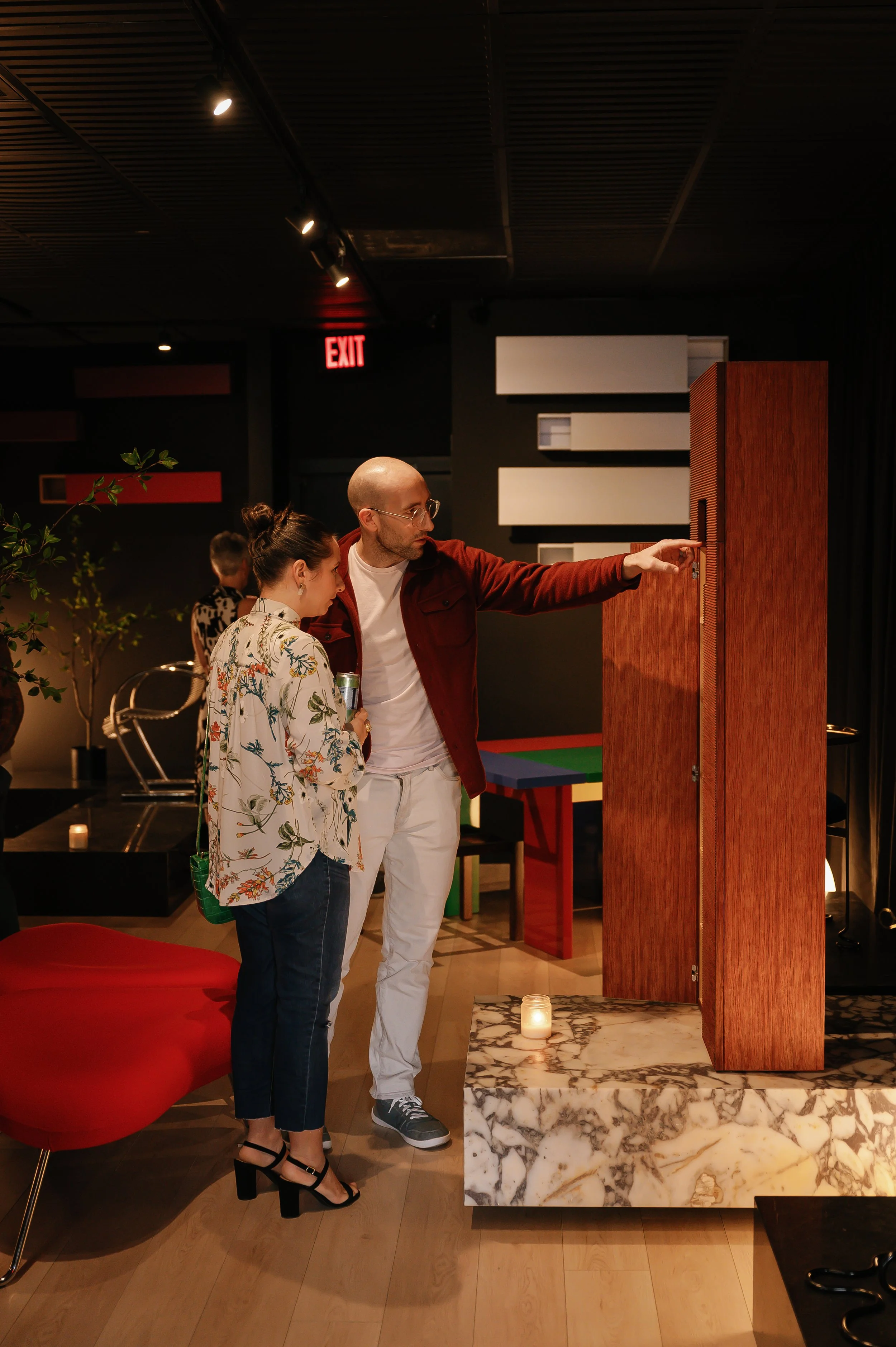Two people, a man and a woman, are in an indoor modern space, examining a tall wooden display or booth. The man is pointing at it while the woman looks on, holding a drink. There is a marble platform and some ambient lighting.