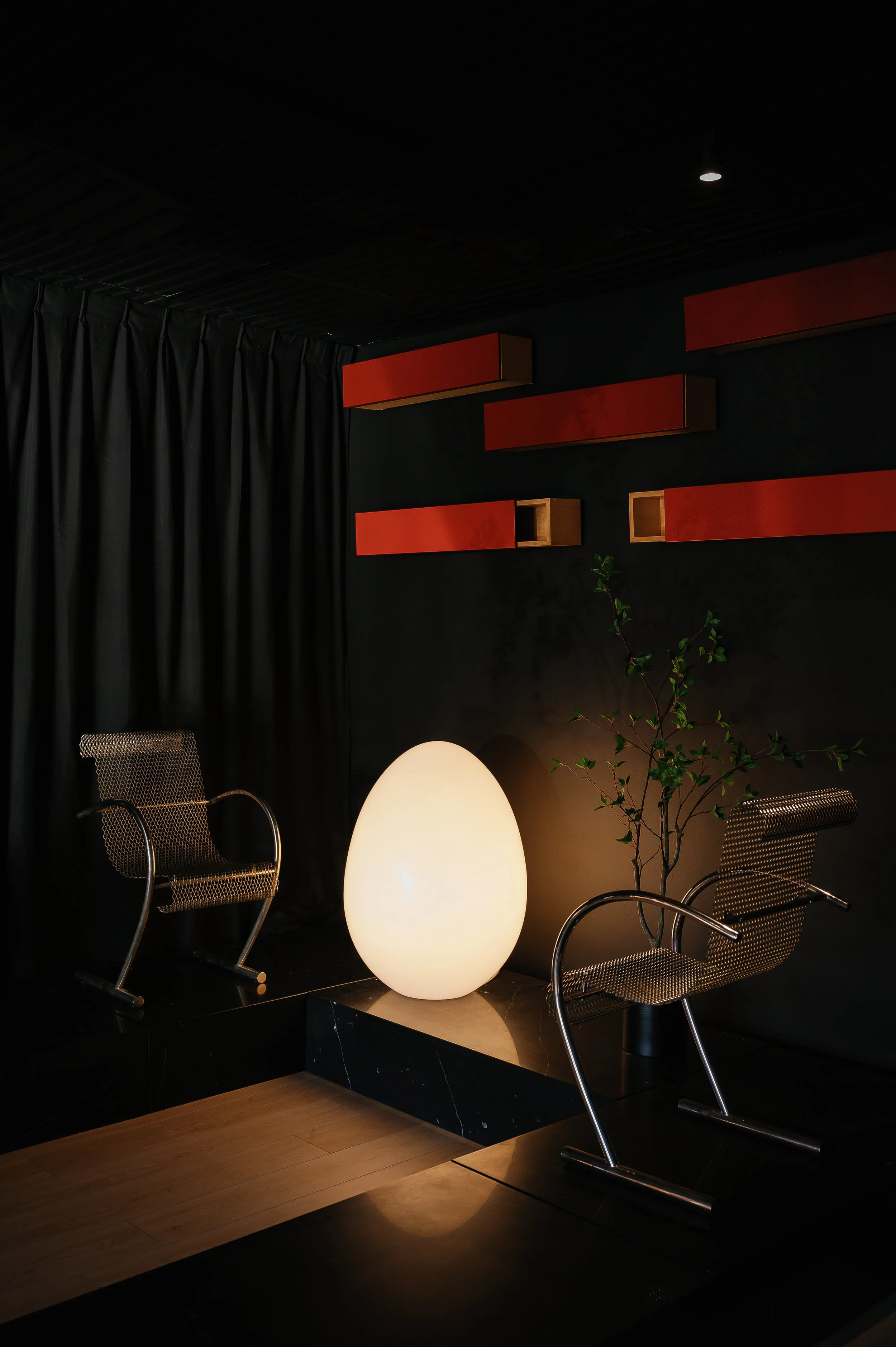 Modern interior with black curtains, red wall-mounted shelves, a large glowing egg-shaped lamp, two metallic chairs, and a small green plant.