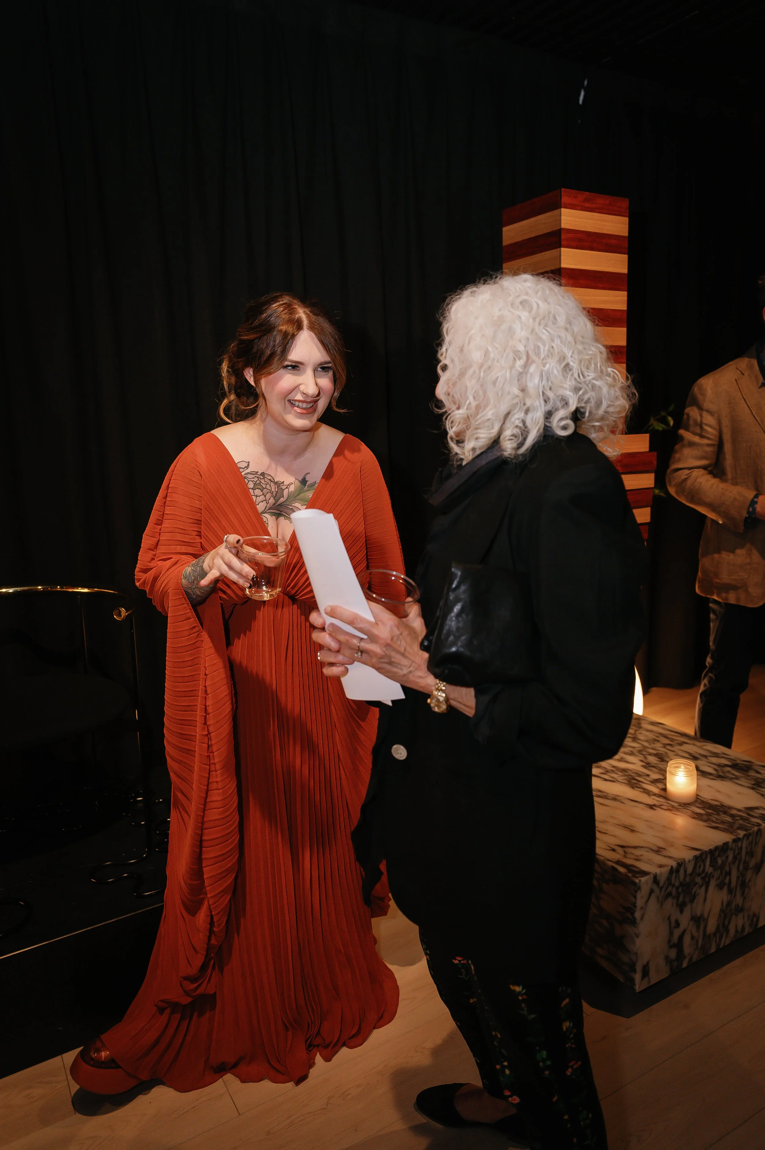 Two women are talking at a social event, holding glasses with drinks. One woman has curly gray hair and is dressed in black, while the other woman has red hair, tattoos, and is wearing an orange dress. They are standing indoors with black curtains an