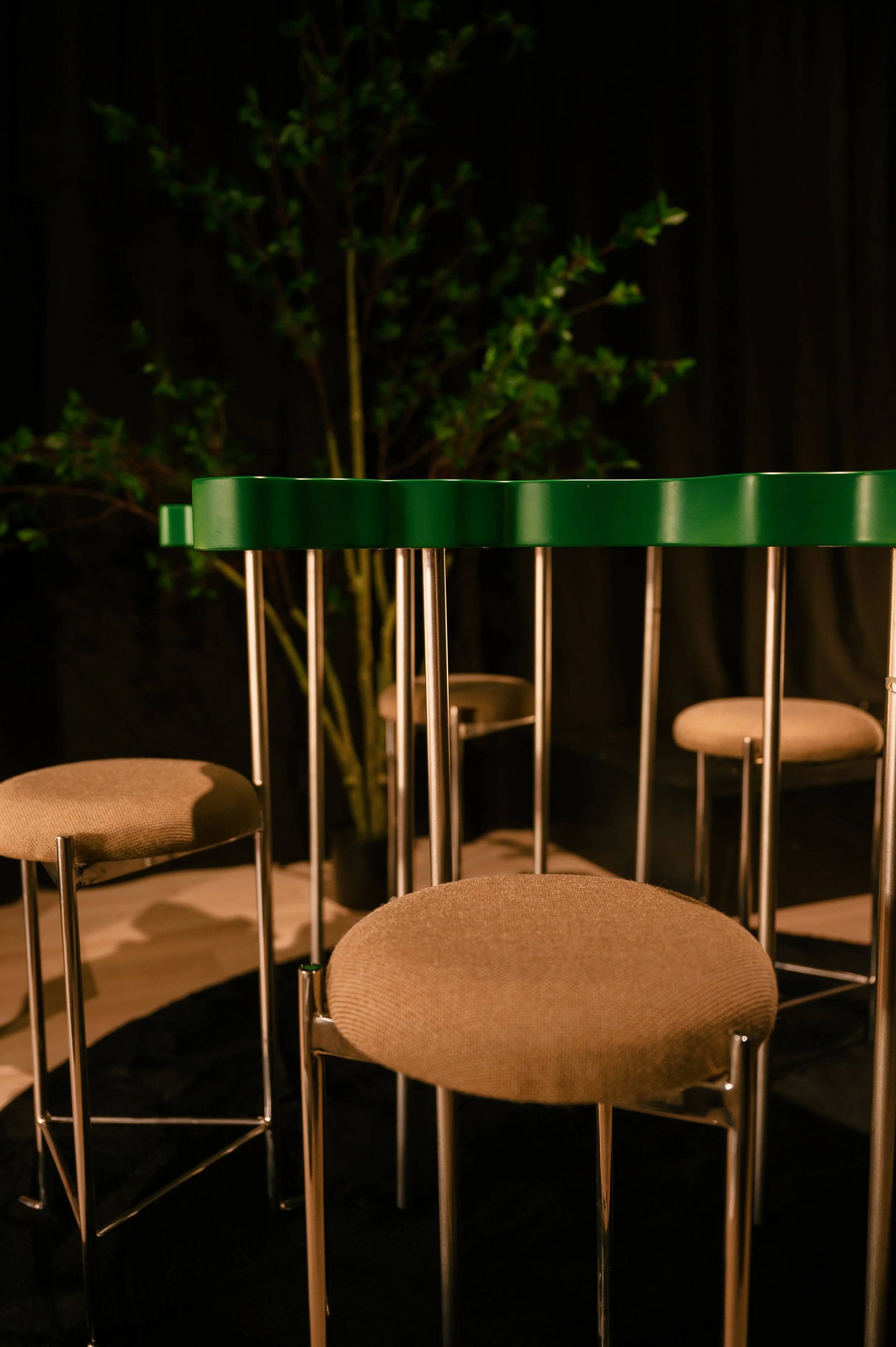 Interior scene with a green high-top table and four beige cushioned stools in front of a black curtain and a potted plant in the background.