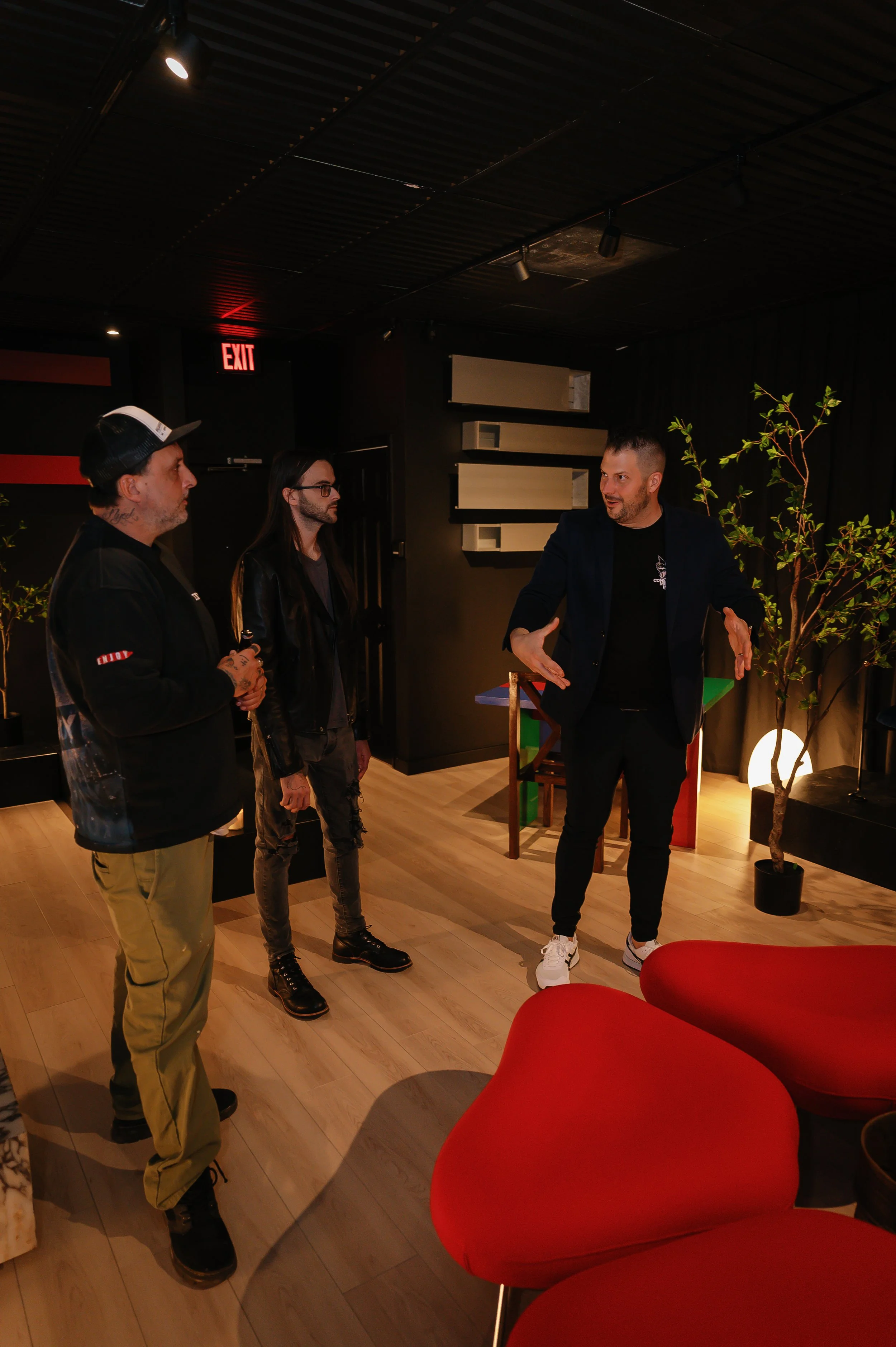 Three men engaged in conversation inside a dimly lit room with modern decor, including a potted plant and red chairs.