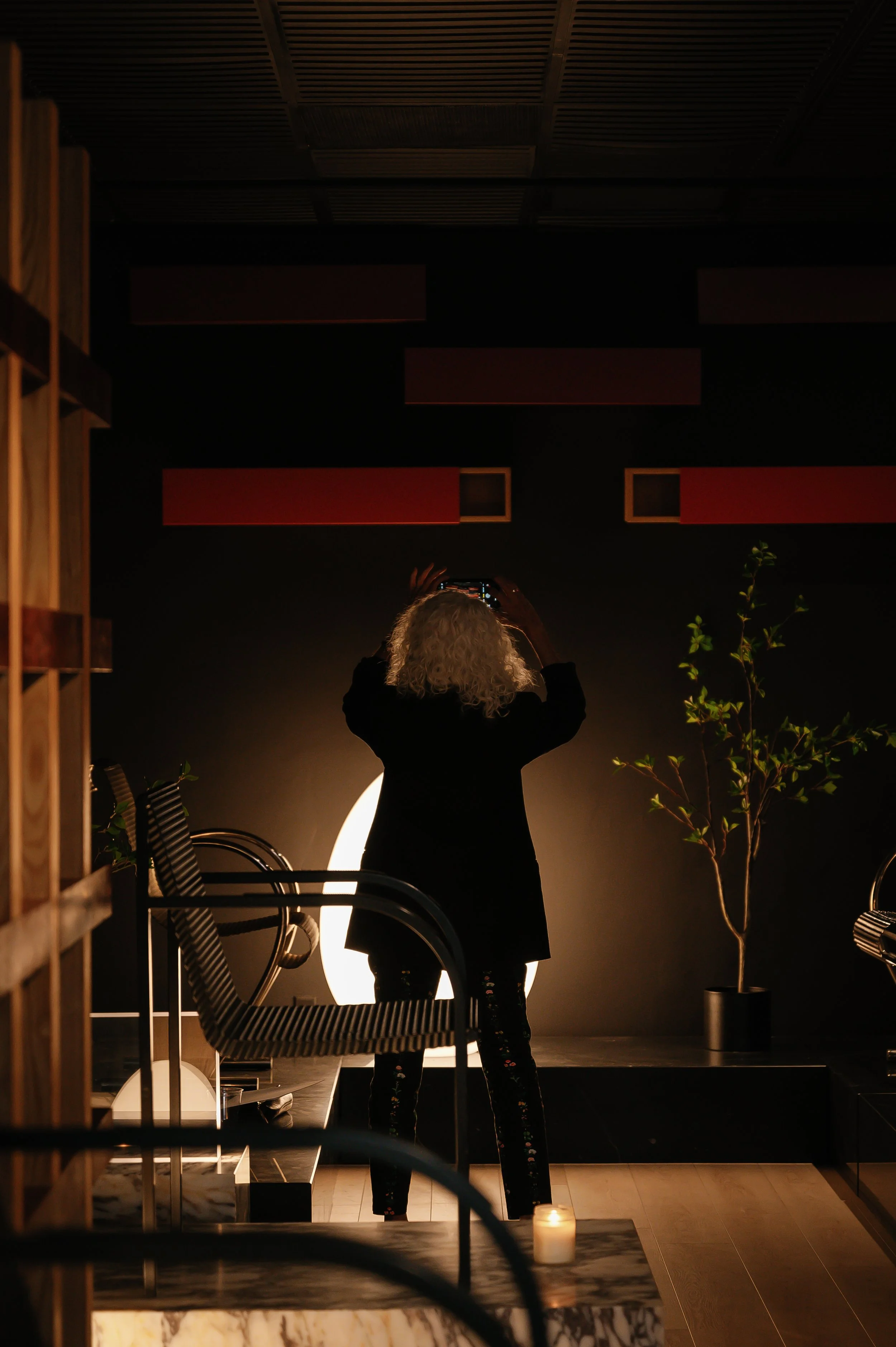 A woman with curly hair takes a photo or selfie in a dimly lit room, standing in front of a bright light source, with a plant and chairs nearby, and a candle on the table.