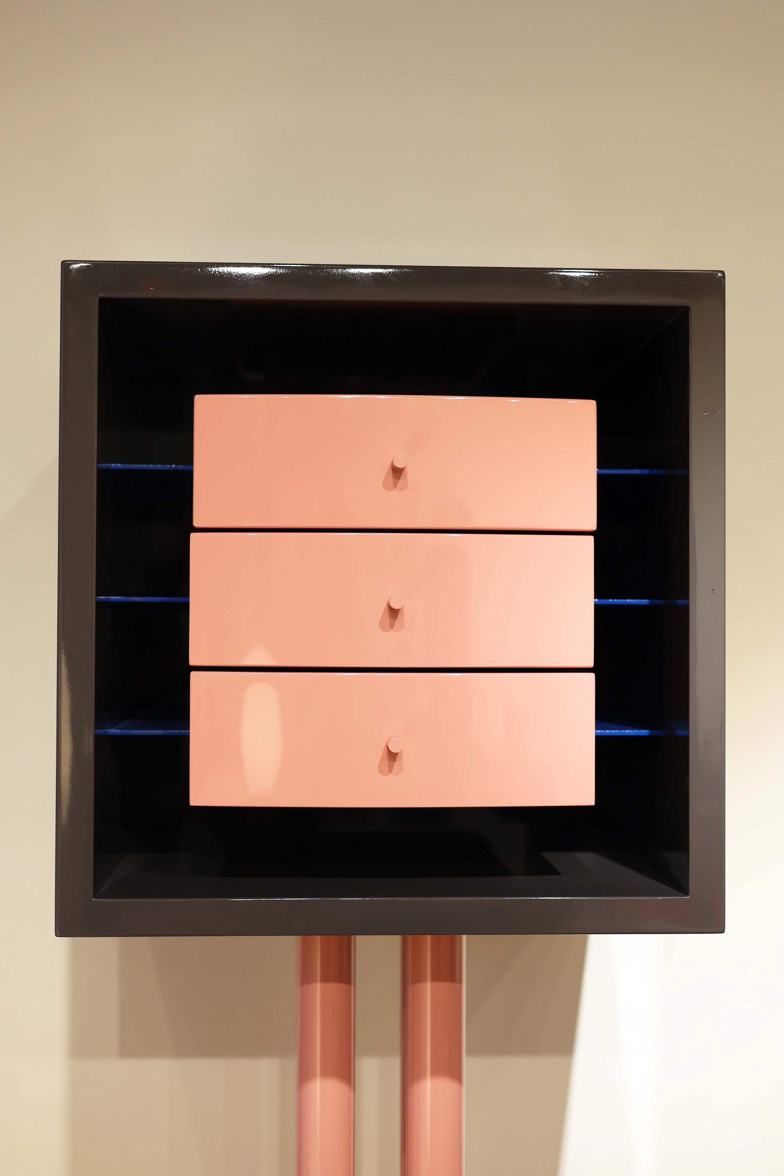 A black square frame with three pink drawers inside, mounted on two pink poles, against a beige wall.