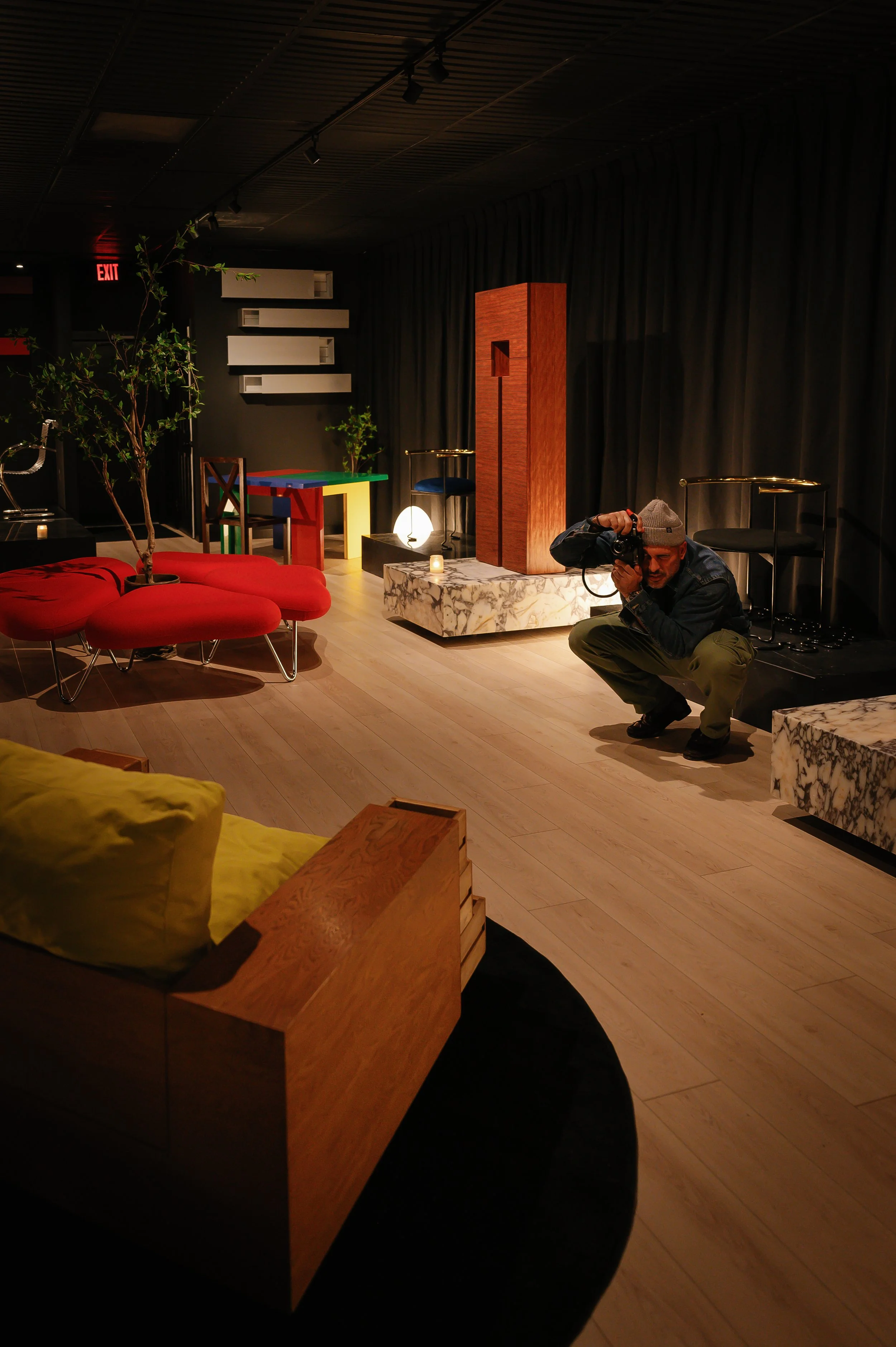 A photographer crouching with a camera in an art gallery with modern furniture, indoor plants, and a dark curtain backdrop.