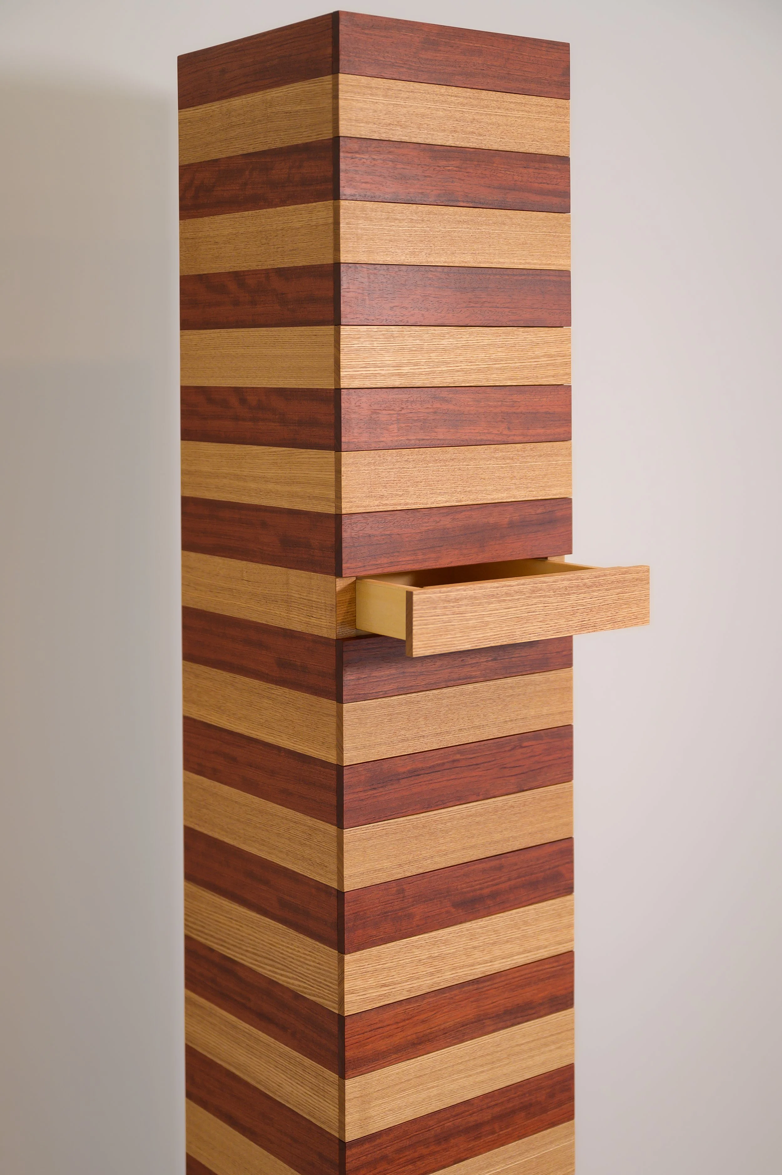 A tall wooden cabinet with horizontal stripes of alternating light and dark wood, partially open drawer in the middle, against a plain wall.