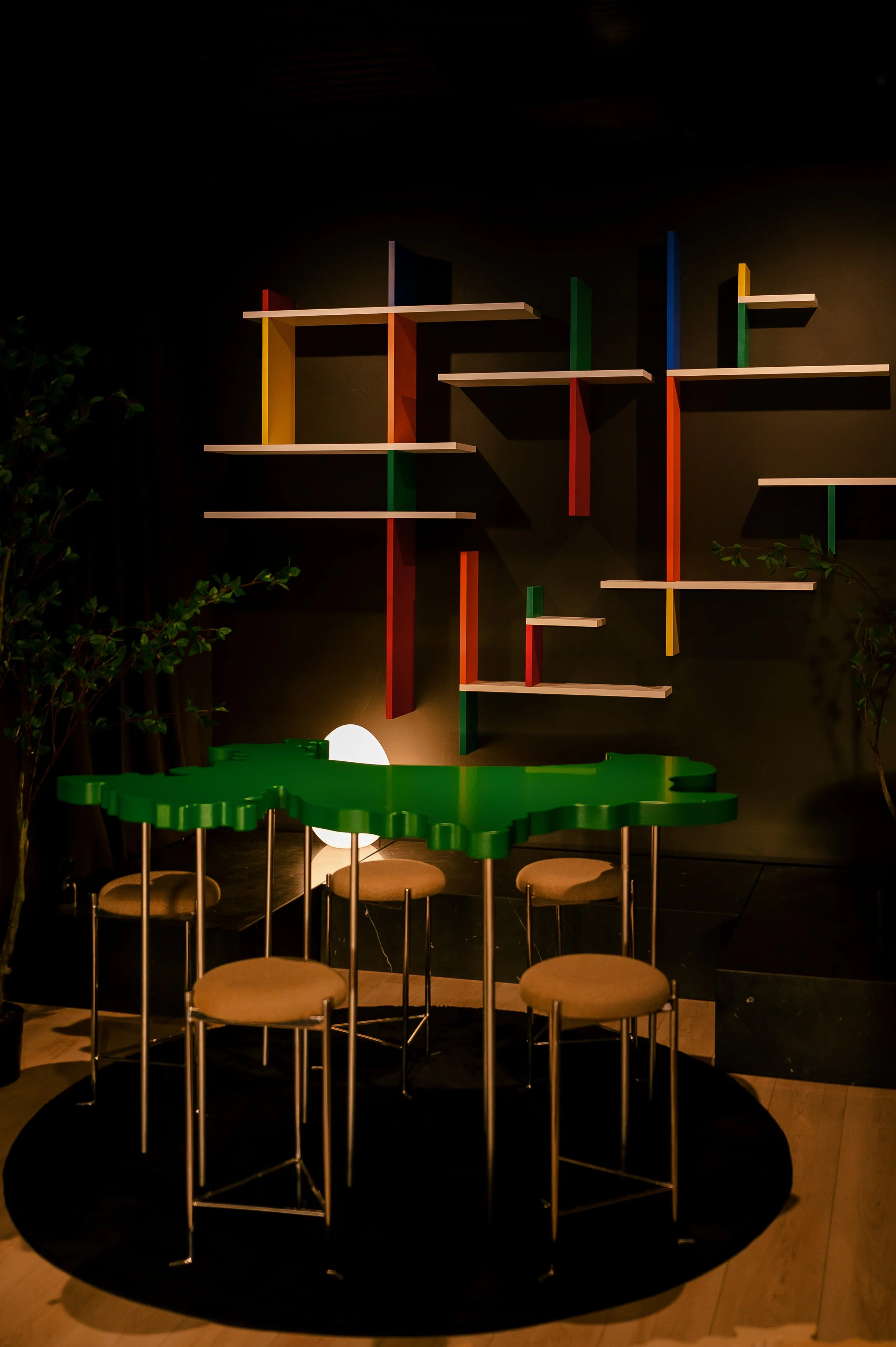 Colorful modern shelves with intersecting horizontal and vertical panels on a dark wall, limited seating with beige cushions around a green irregularly shaped table, dim lighting, potted plants nearby.