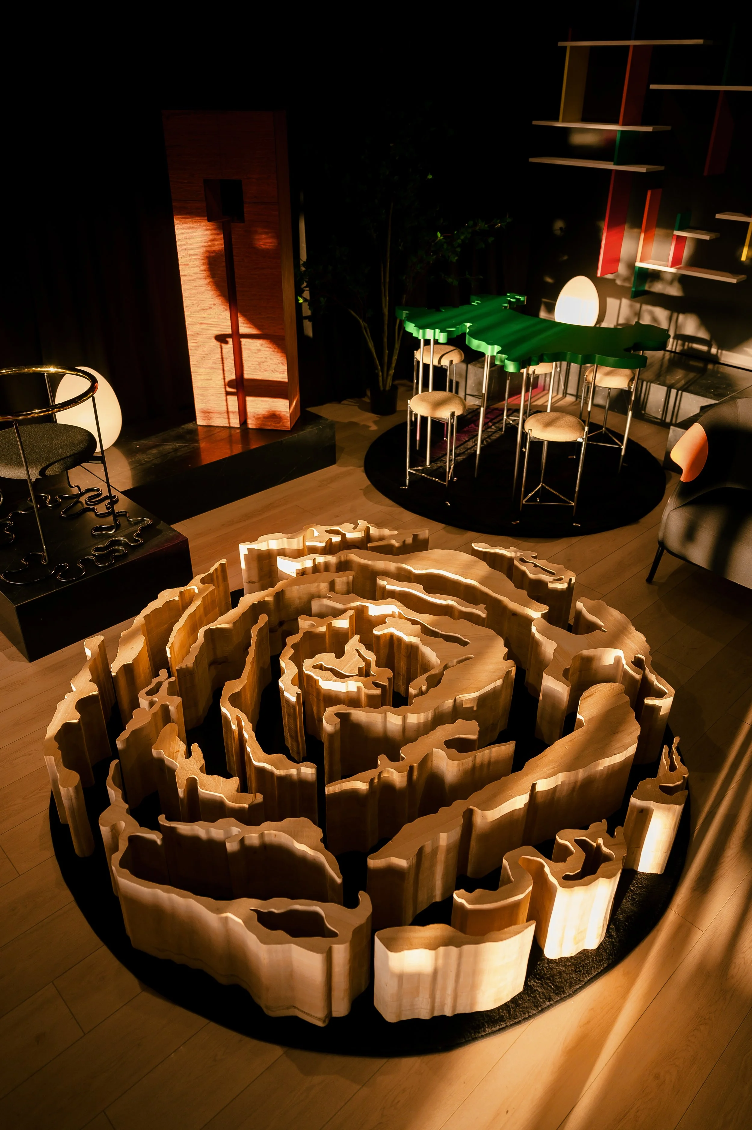Art installation of a large wooden spiral sculpture displayed on a circular black rug in a dimly lit room.