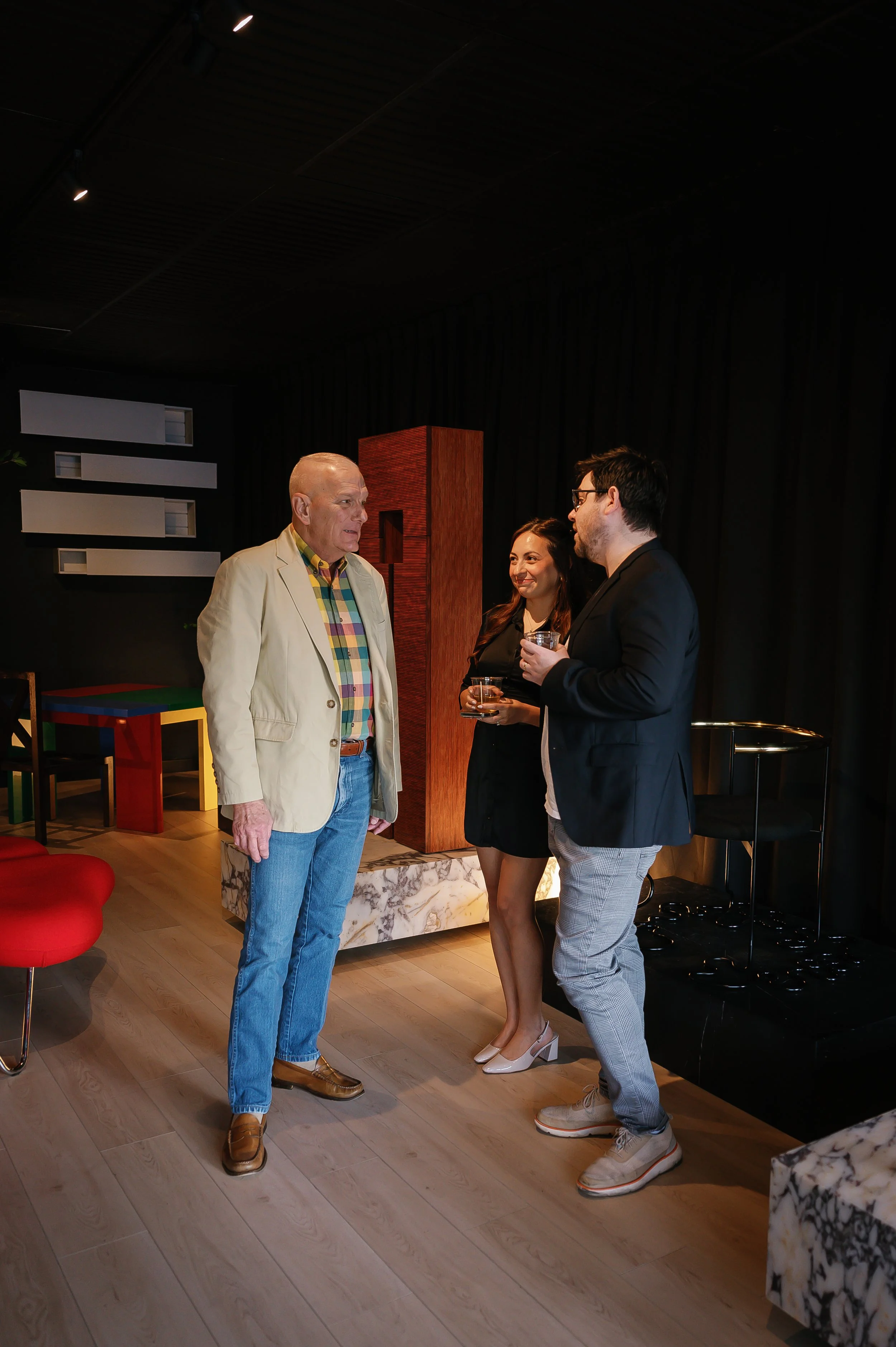 Three people having a conversation at a social gathering, two men and a woman, holding drinks in a modern room with colorful furniture and dark curtains.