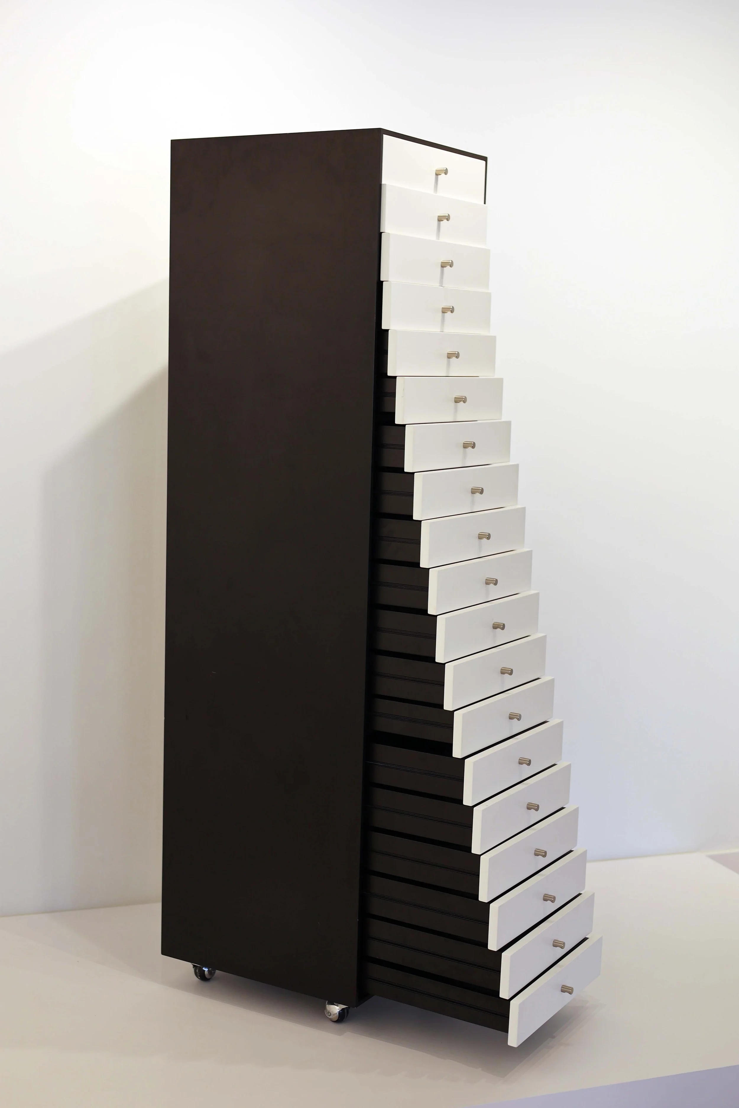 Tall black cabinet with 17 white drawers, some partially open, on caster wheels in a minimal white room.