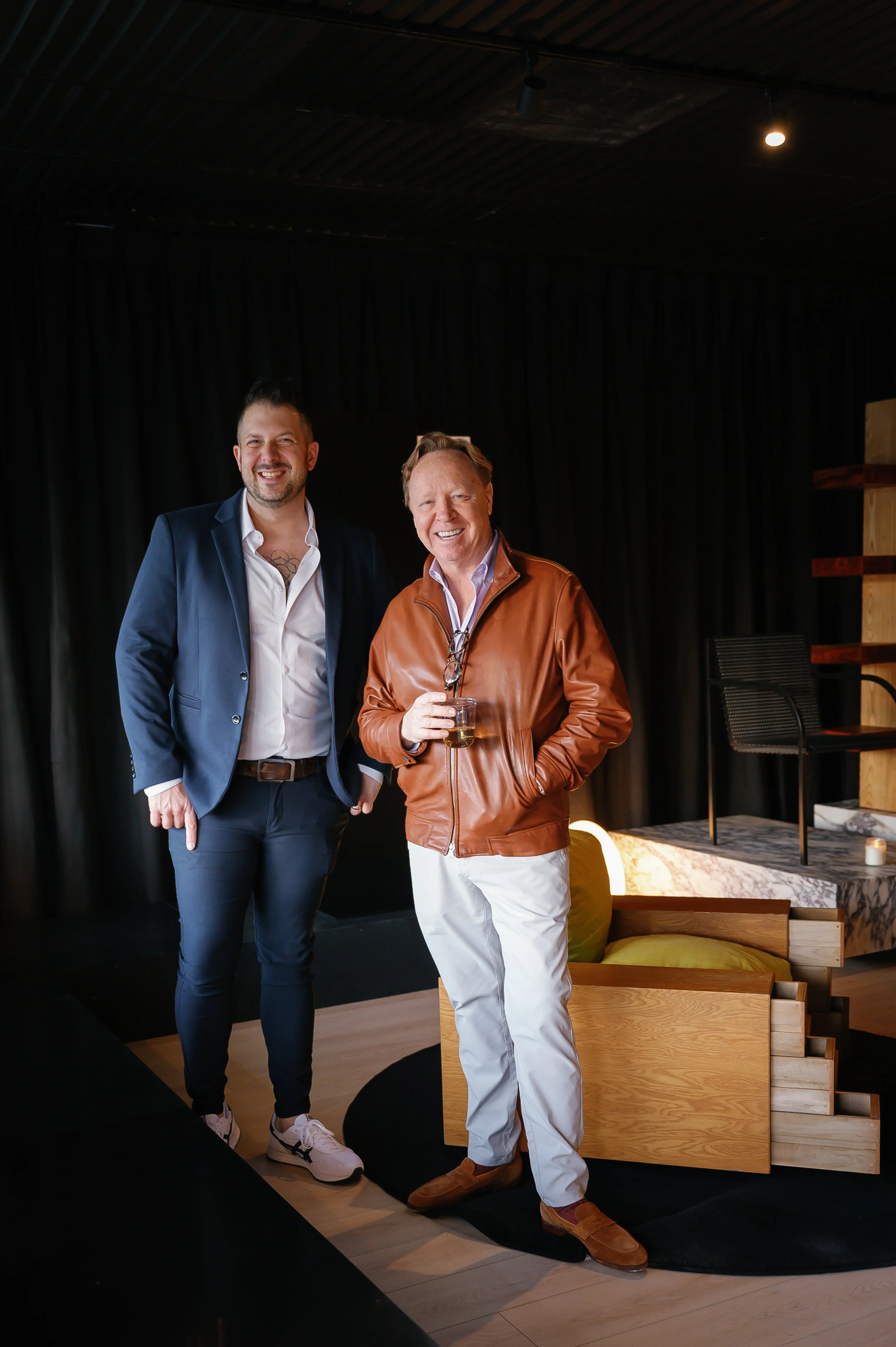 Two men smiling and standing together in a modern stylish room, one holding a drink, with black curtains and contemporary furniture in the background.