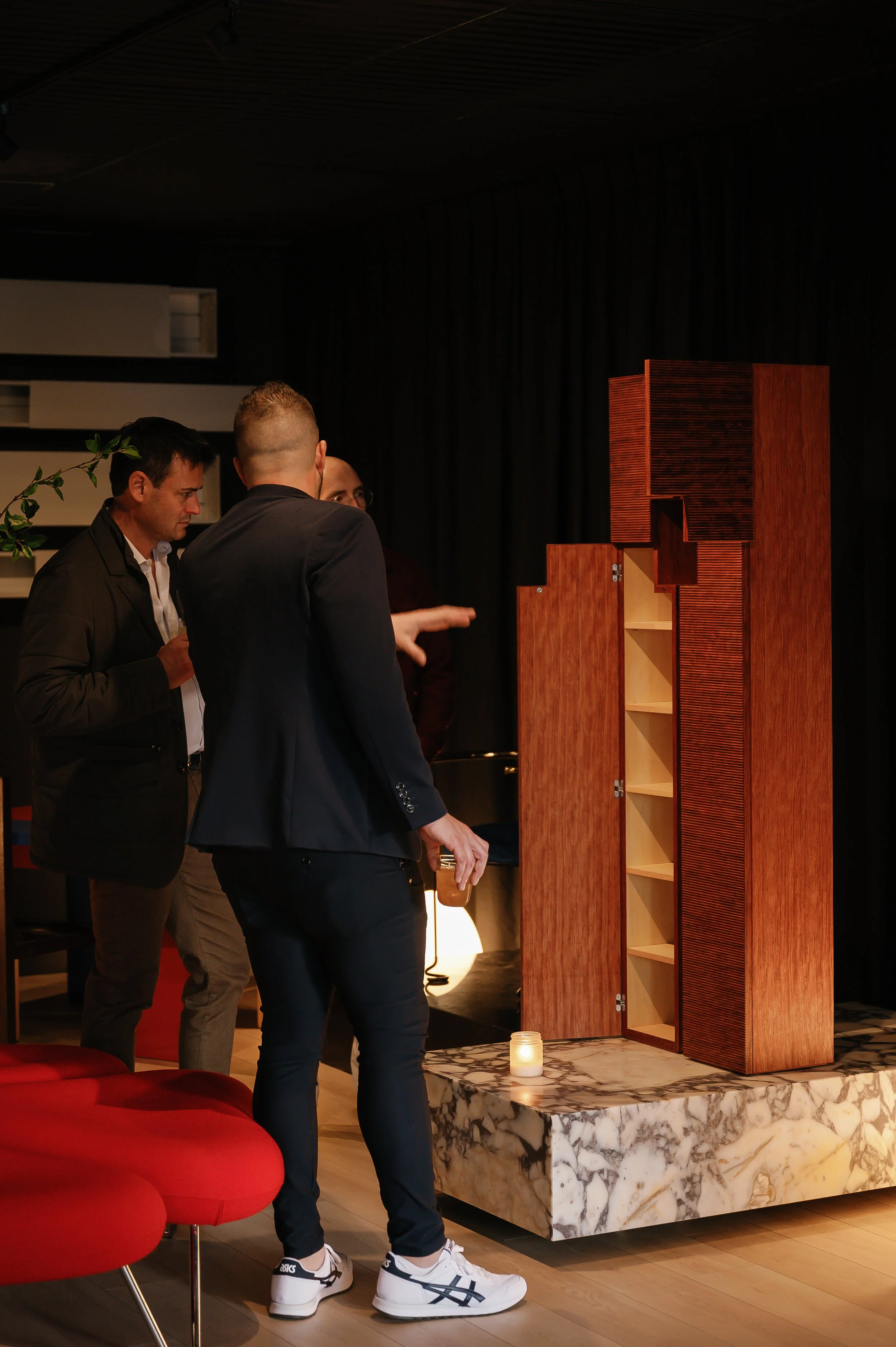 Three men are looking at a wooden cabinet with open doors and shelves, displayed on a marble platform, in a dimly lit indoor setting.