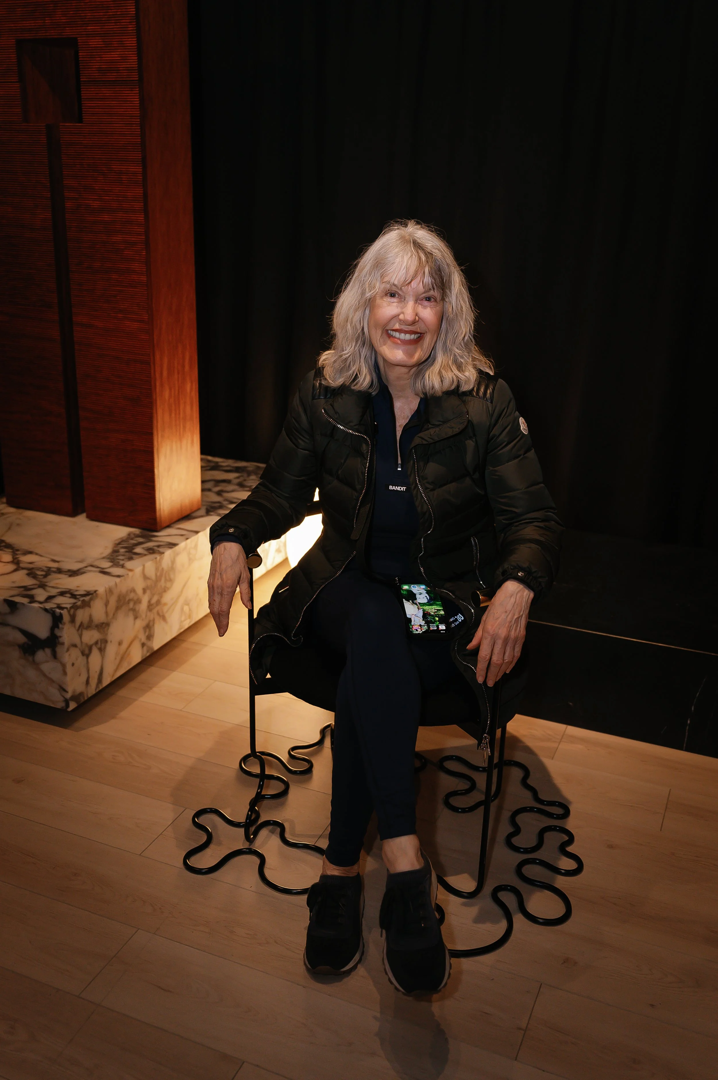 A smiling woman with blonde, wavy hair sitting on a black chair inside a room with wood and black decor. She is wearing a black jacket, dark pants, and black sneakers, and holding a smartphone. There is a marble platform and black curtains in the bac