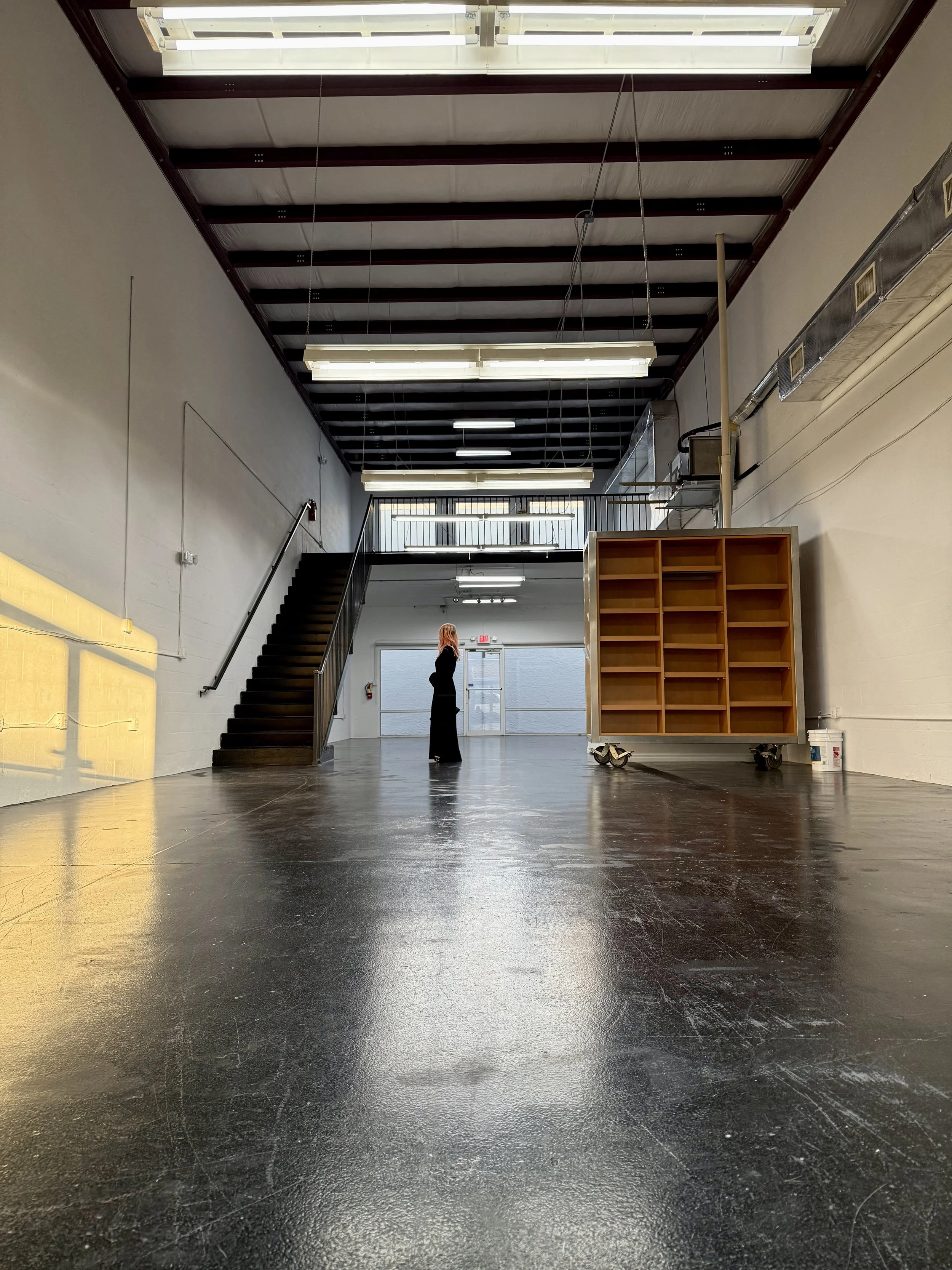 An empty industrial-style space with black polished floors, white walls, exposed ceiling beams, and fluorescent lighting. A woman stands near a staircase by a glass door, with a large empty wooden shelving unit on wheels nearby.